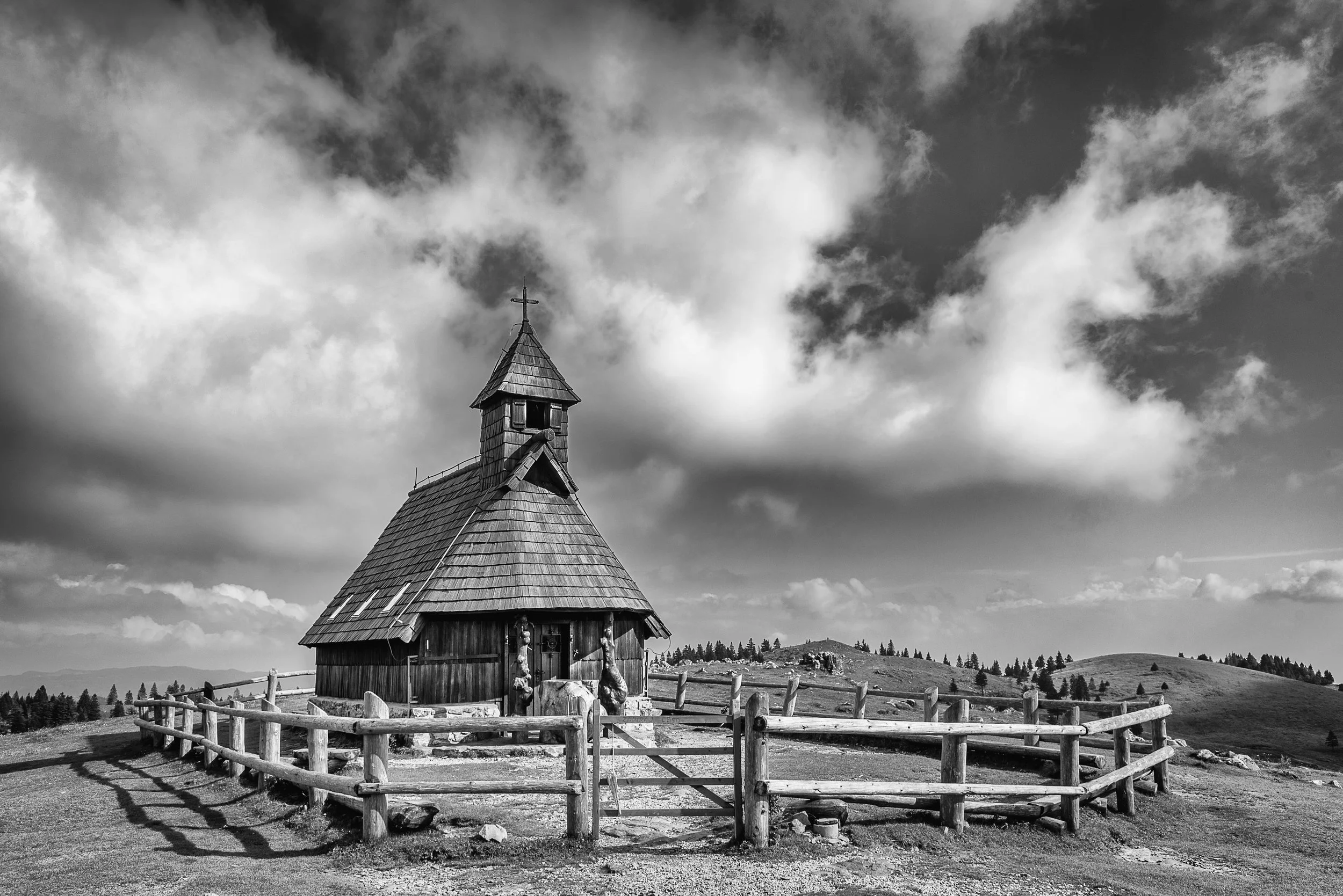 Black and white photo of a small wooden church with a fence around it, set on a hill under a cloudy sky.