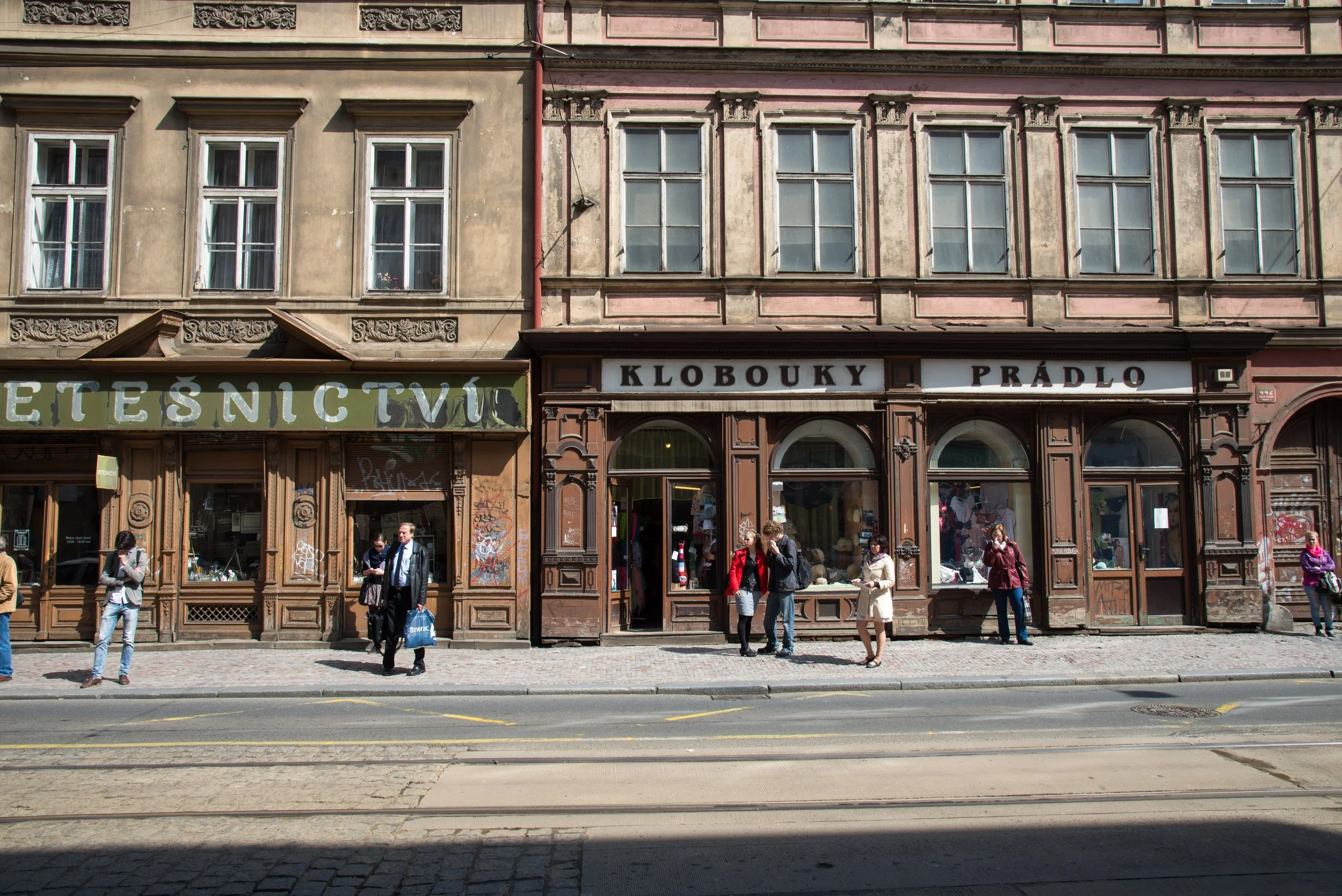 People standing on the sidewalk in front of a historic building with storefronts, some engaged in conversation.
The building has a faded beige upper facade and ornate wooden framing around the storefronts on the ground level. There are signs in a for