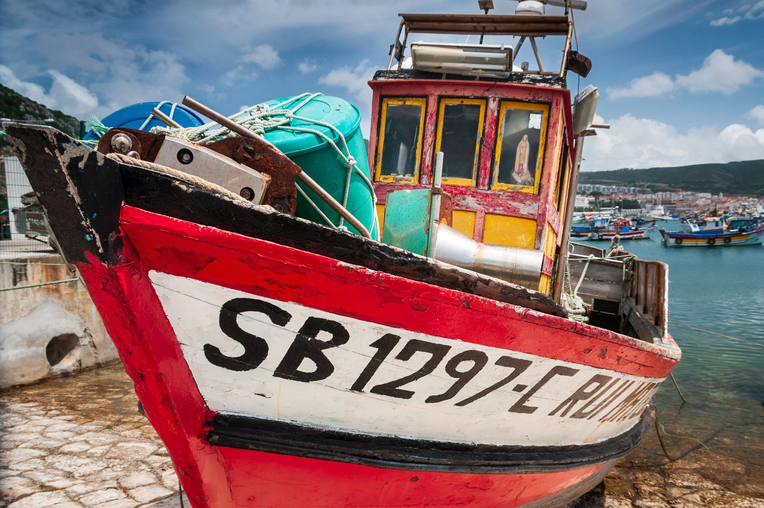 Colorful fishing boat docked at a harbor with many other boats in the background, under a partly cloudy sky.