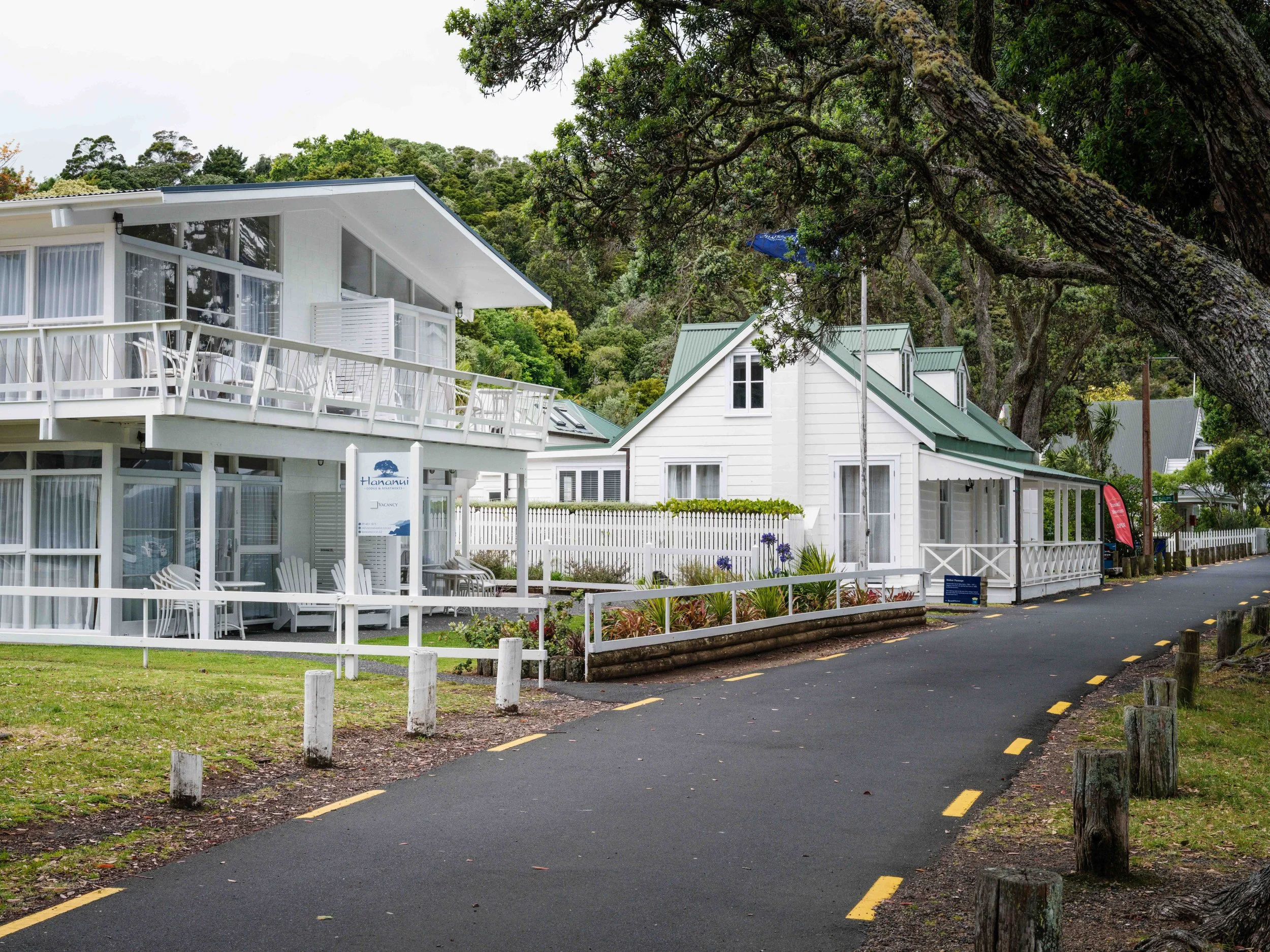 White residential houses with green roofs alongside a paved walking path, surrounded by large trees and greenery.