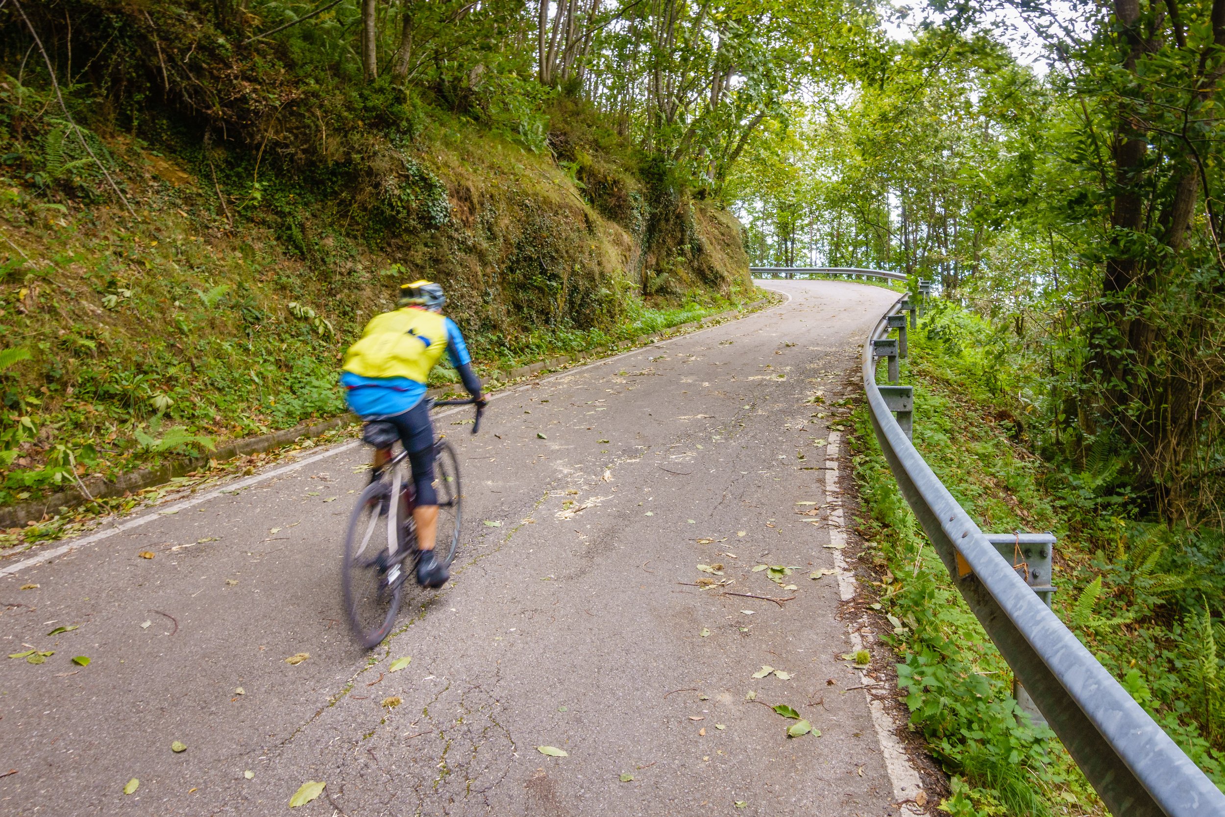 A cyclist in a yellow jacket riding on a winding mountain road surrounded by green trees and foliage.