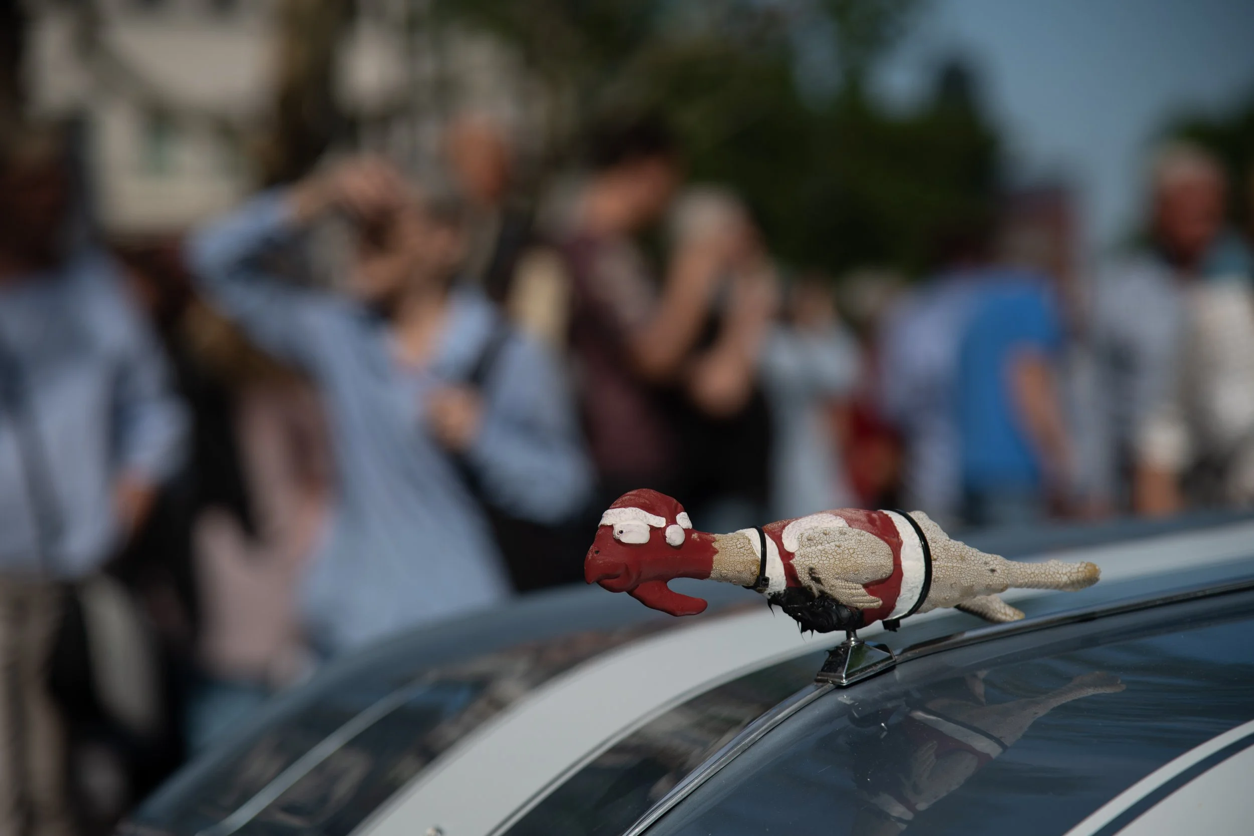 A small dachshund-shaped ornament dressed in a Santa suit on top of a police car with a crowd of people looking and taking photos in the background.