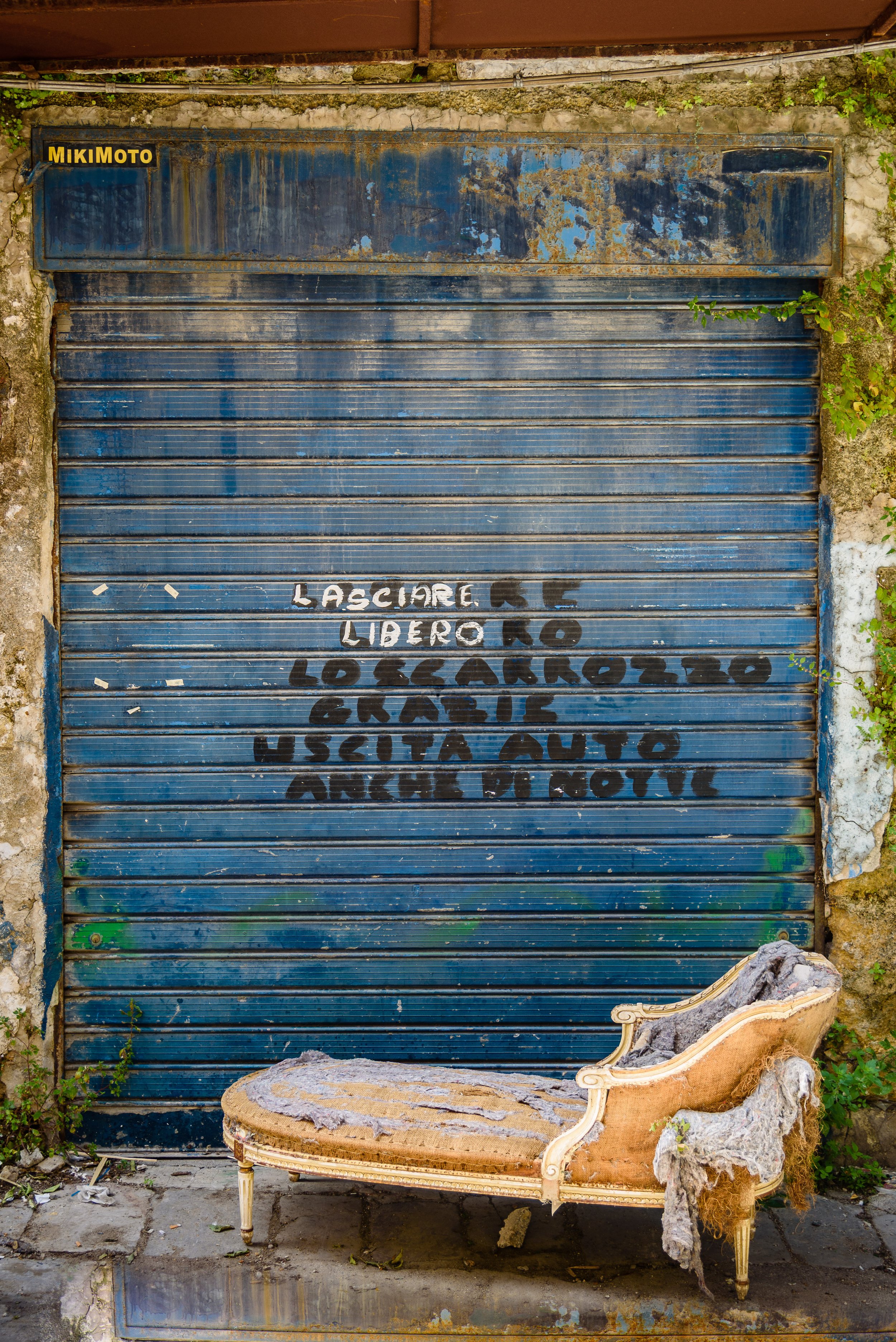 Old, weathered blue metal garage door with a yellow and black sign that says 'MIKI MOTO' in the top left corner. Graffiti painted on the door in white and black includes Italian phrases such as 'LASCIARE LIBERO,' 'LO SCARROZZO,' and 'USETA AUTO ANGUI