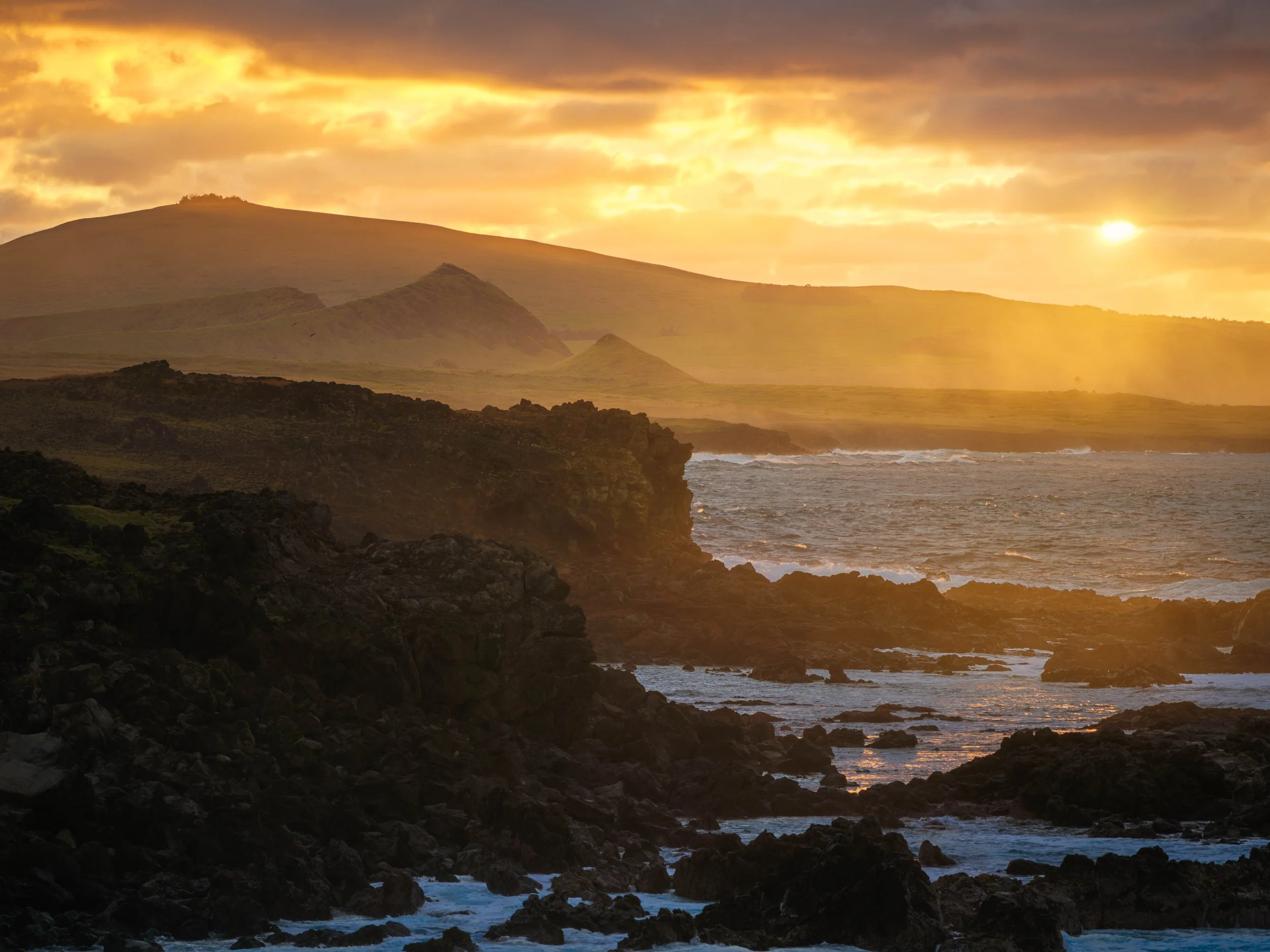 Sunset over a rugged coastline with dark rocks, waves, and a misty grassy hill in the background