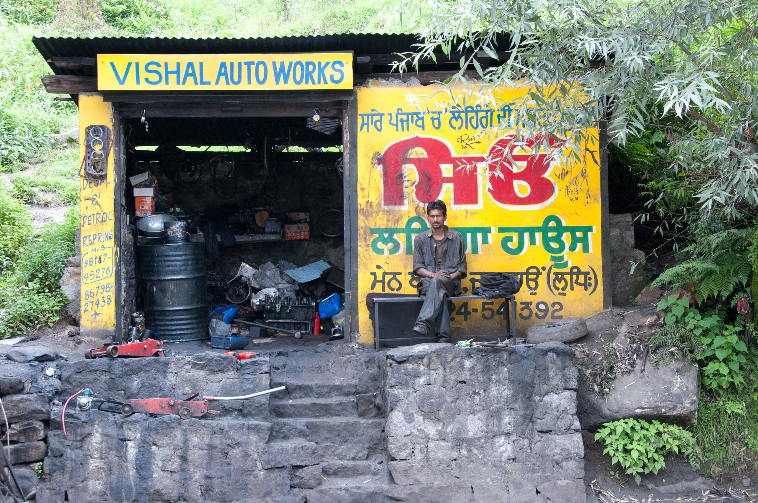 A man sitting on a bench outside an auto repair shop with a yellow sign that reads 'VISHAL AUTO WORKS,' surrounded by green foliage and tools, with a partially open garage showing car parts and equipment inside.