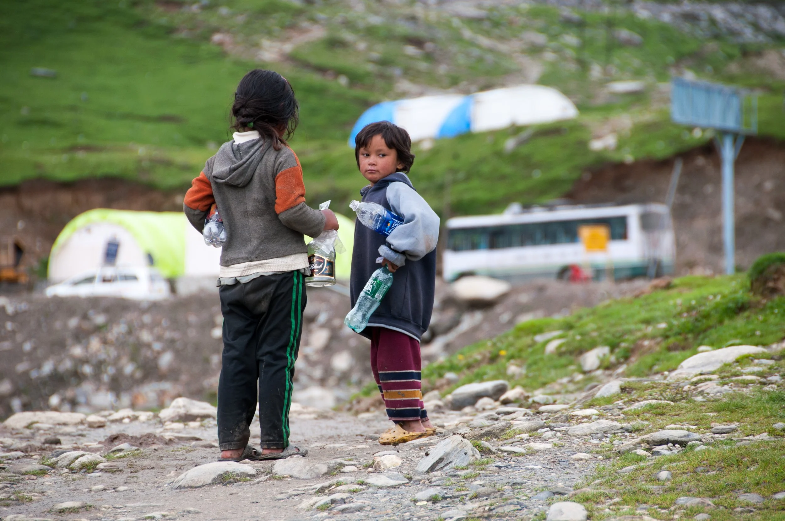 Two children standing on a rocky path outdoors, holding water bottles and talking, with huts and a bus in the background.