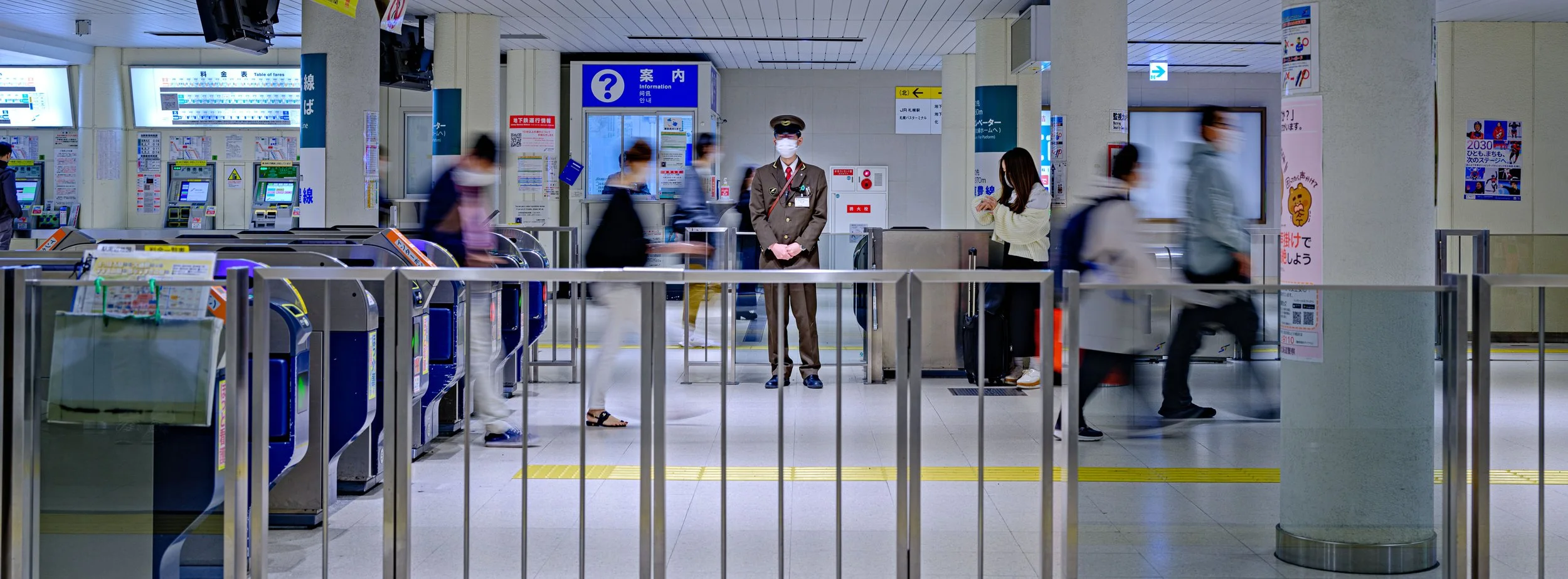 People walking through a security checkpoint at a train station or airport, with a station staff member in uniform standing in the middle, and electronic ticket gates on the side.