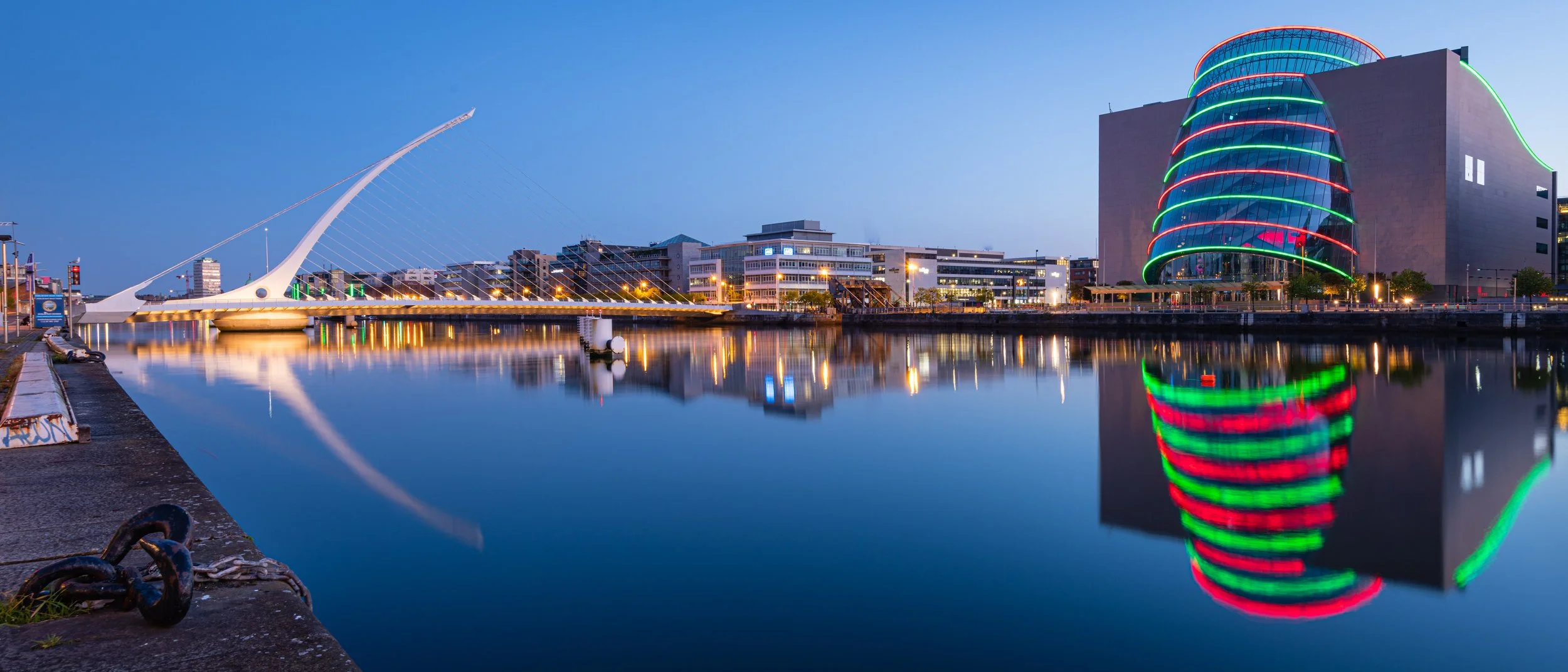 City skyline with modern buildings and a bridge along a calm river at dusk, city lights reflecting on the water, with a distinctive curved glass building decorated with colorful neon lights.