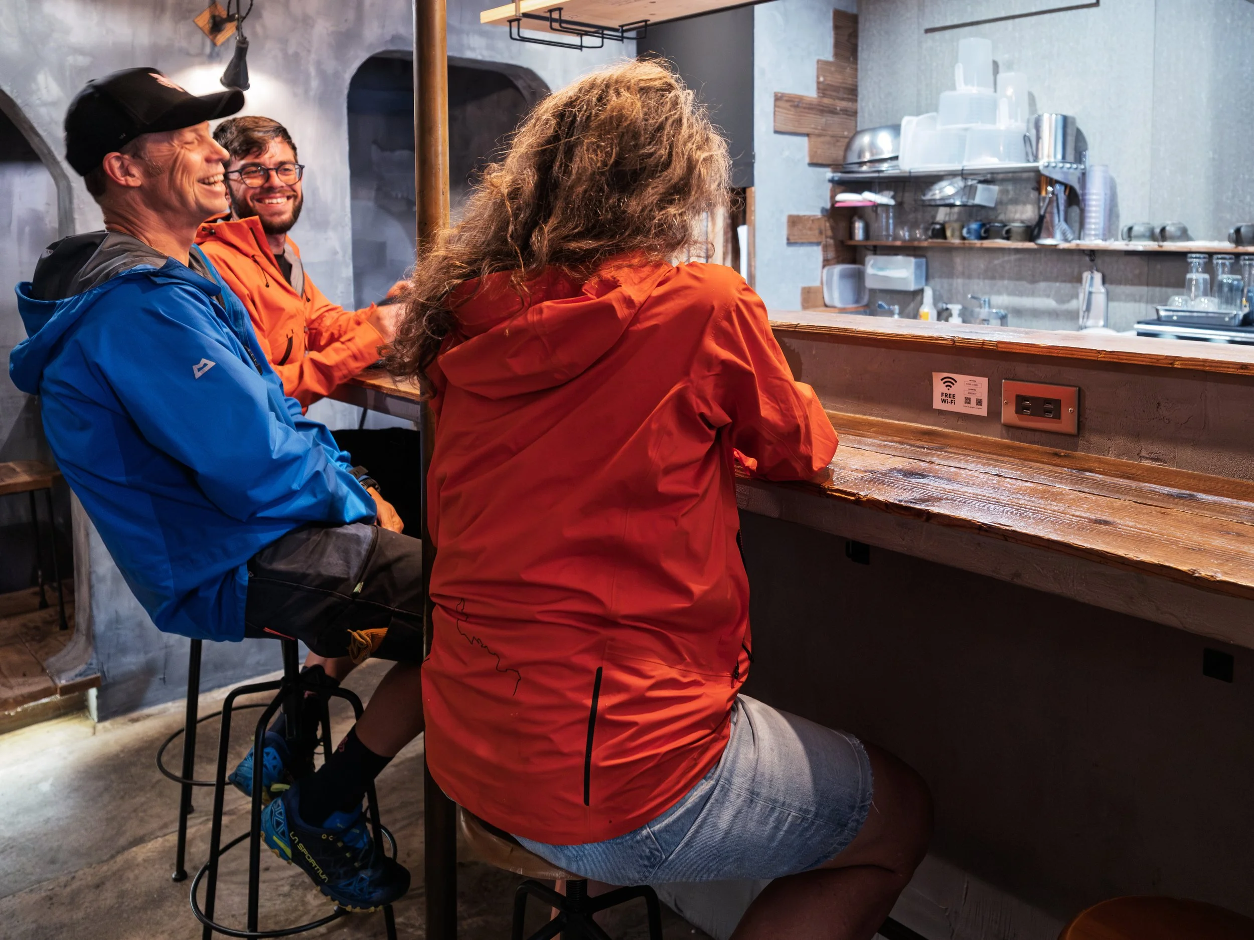 Three people sitting at a bar counter in a cozy cafe. One woman facing away, with long curly hair, wearing a bright orange jacket. Two men smiling, one in a blue jacket and cap, the other in an orange jacket with glasses. The cafe has a rustic interi
