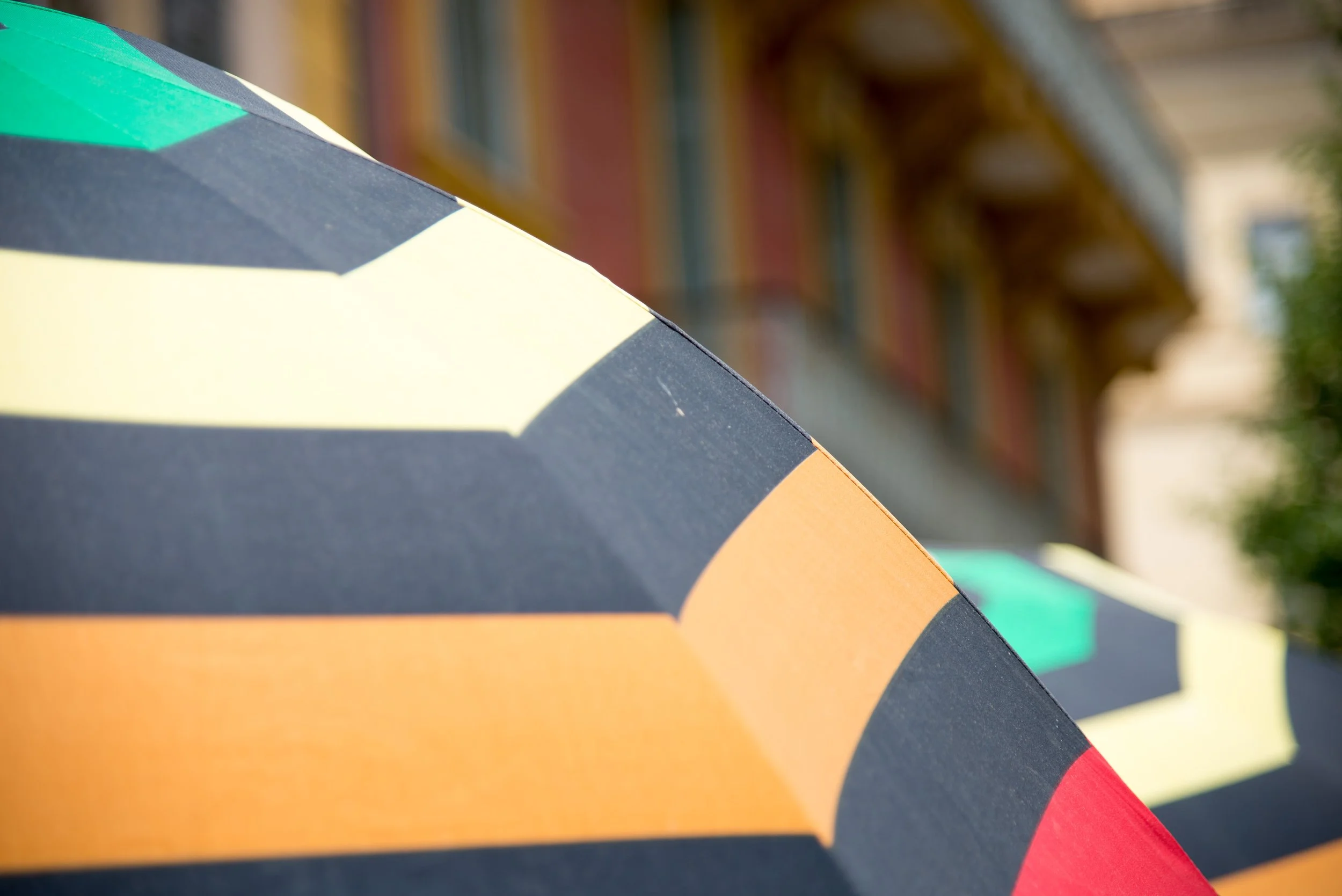 Close-up of a colorful umbrella with orange, yellow, and teal sections, outdoors with a blurred building in the background.