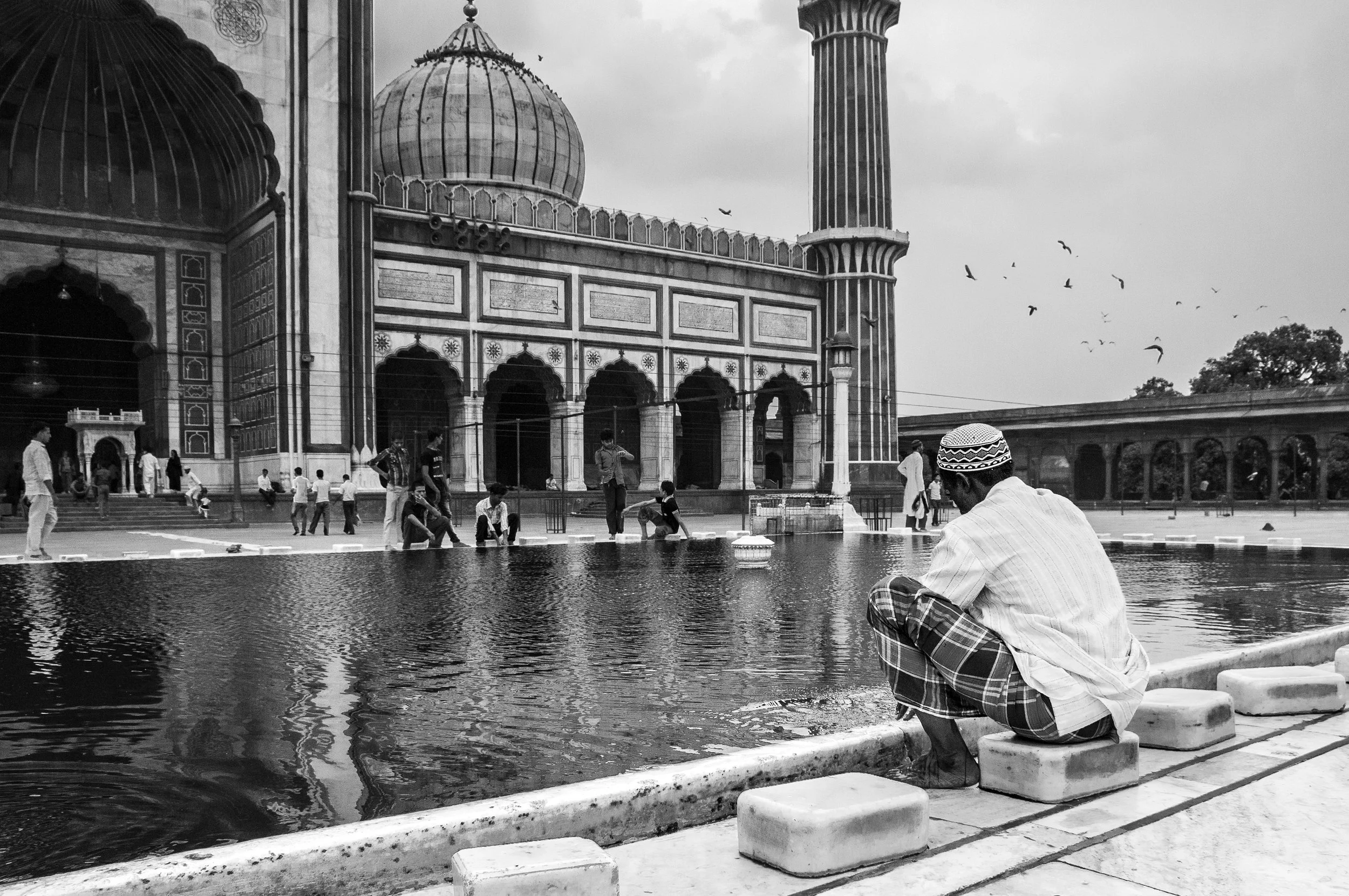 A man in traditional clothing, sitting by a water feature in front of the Jama Masjid mosque in India, with people walking and birds flying in the background.