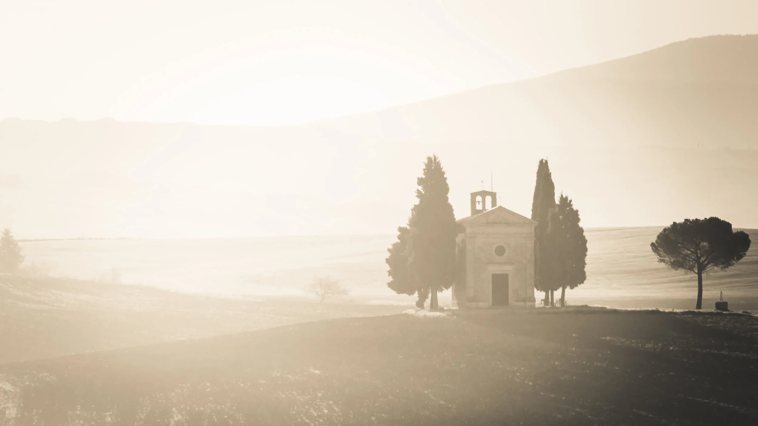 A small church surrounded by tall cypress trees in a misty, rural landscape with rolling hills in the background.