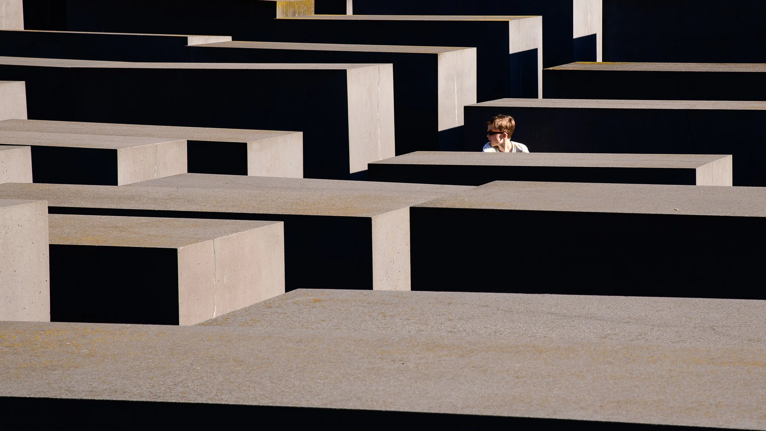 A young boy wearing sunglasses and a white shirt leaning on a concrete slab in a memorial with numerous large concrete blocks creating a geometric pattern.