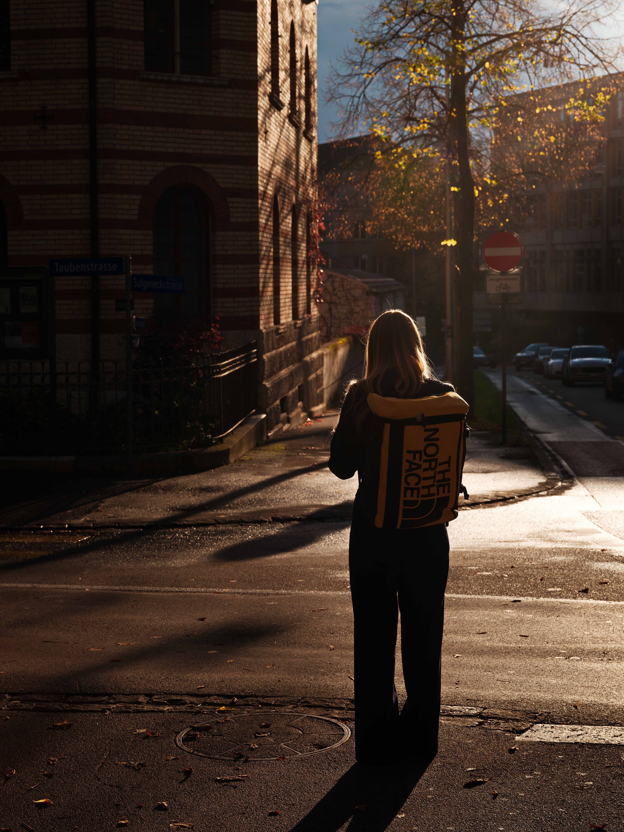 A person with long hair wearing a backpack walking on a street during sunset, casting a long shadow, with trees and buildings in the background.
