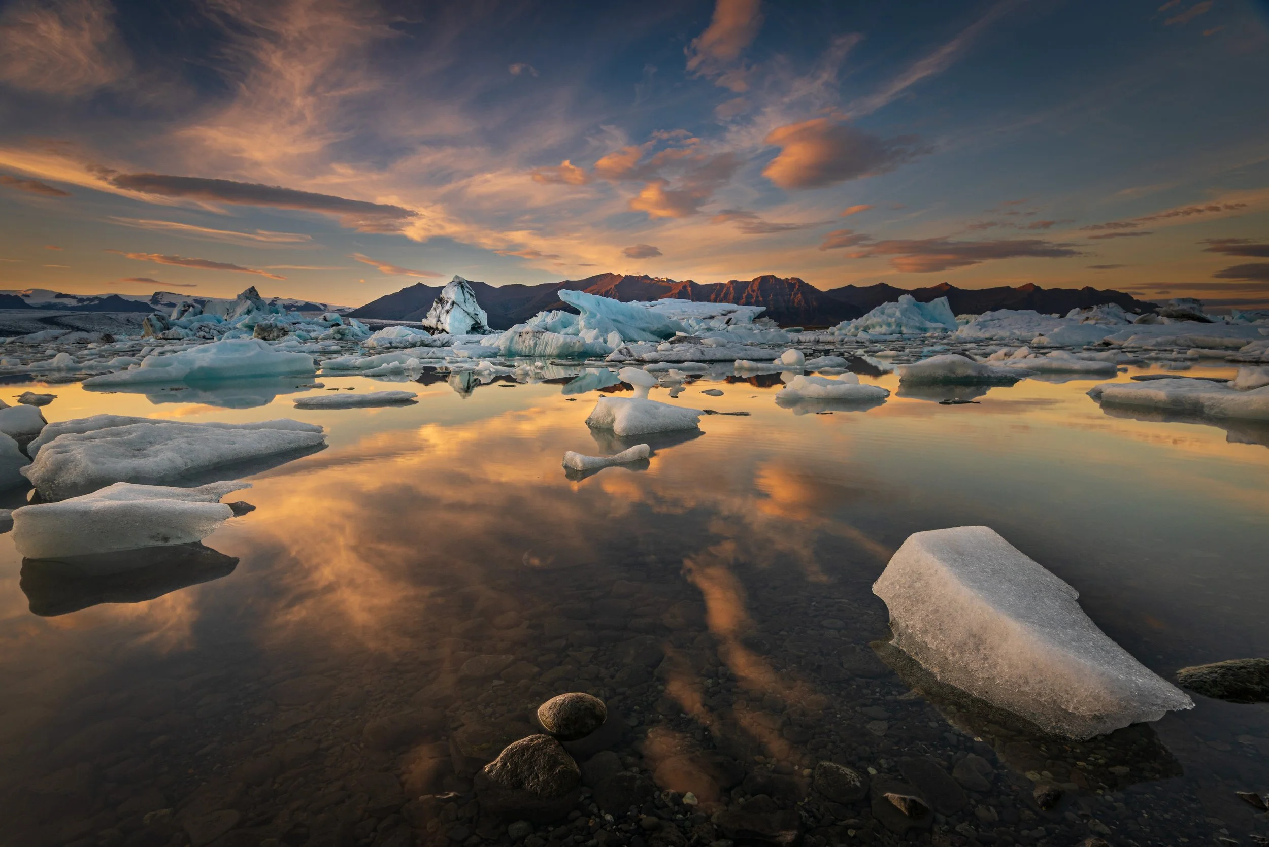 Icebergs floating in a calm body of water during sunset with mountains in the background and a partly cloudy sky.