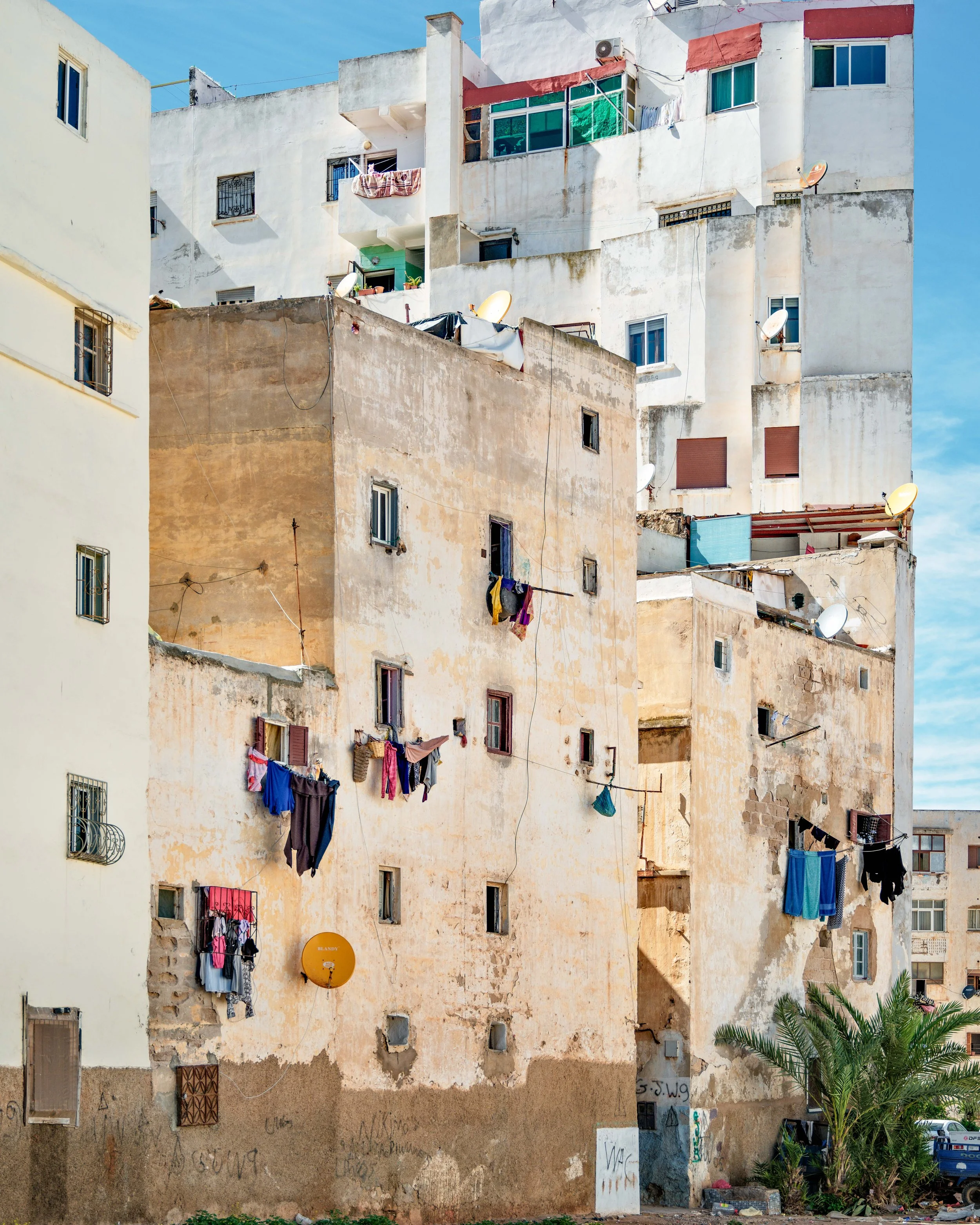 A multi-story residential building with a worn, weathered facade, laundry hanging outside the windows, and a palm tree in front under a clear blue sky.