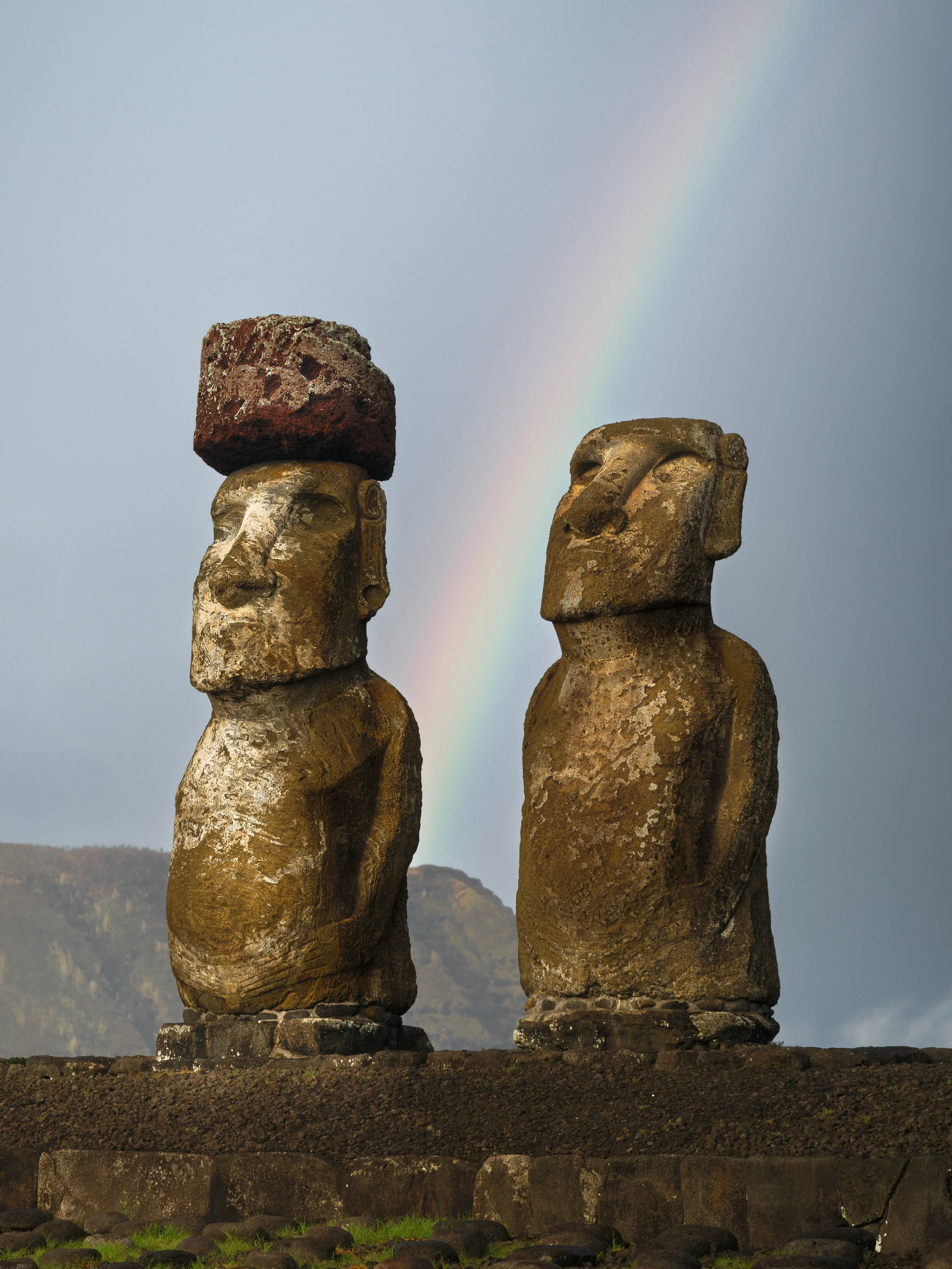 Two Moai statues with a rainbow in the sky behind them.