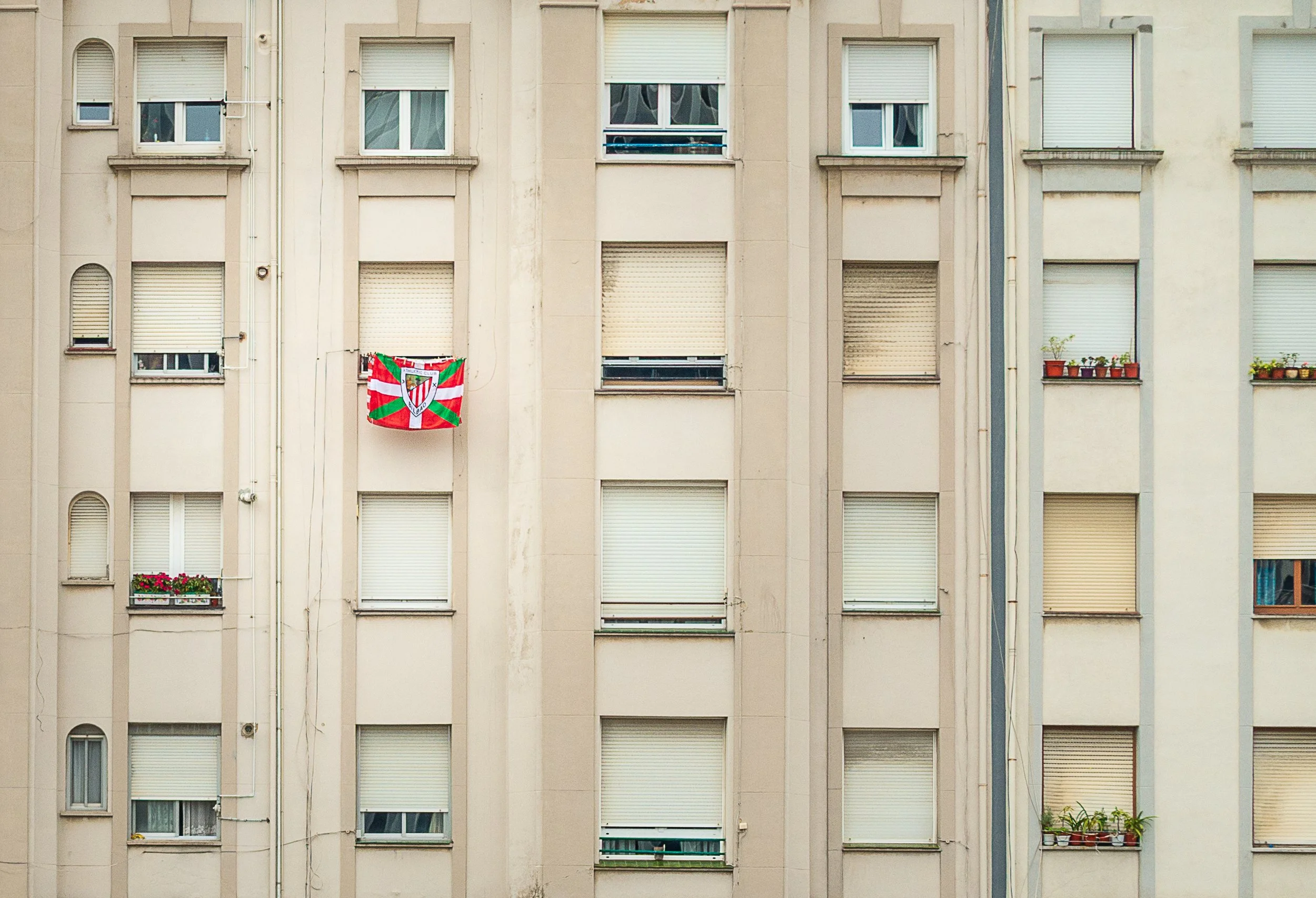 Close-up of a beige apartment building with several windows, some with open blinds and flower pots, and a colorful flag hanging on the second floor from the left.