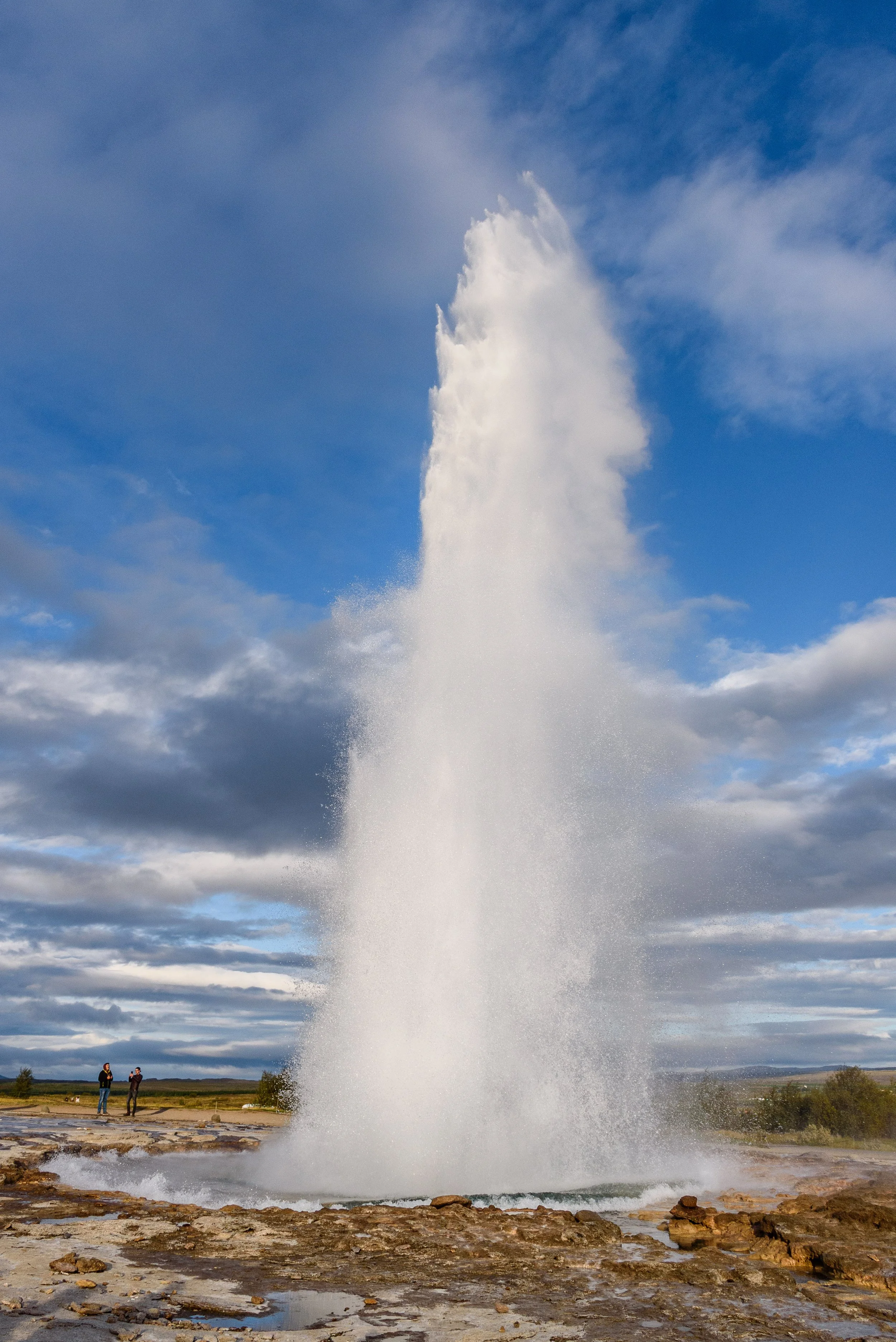 A tall geyser erupting with water shooting high into the sky in Iceland, with a partly cloudy blue sky and two people observing in the background.