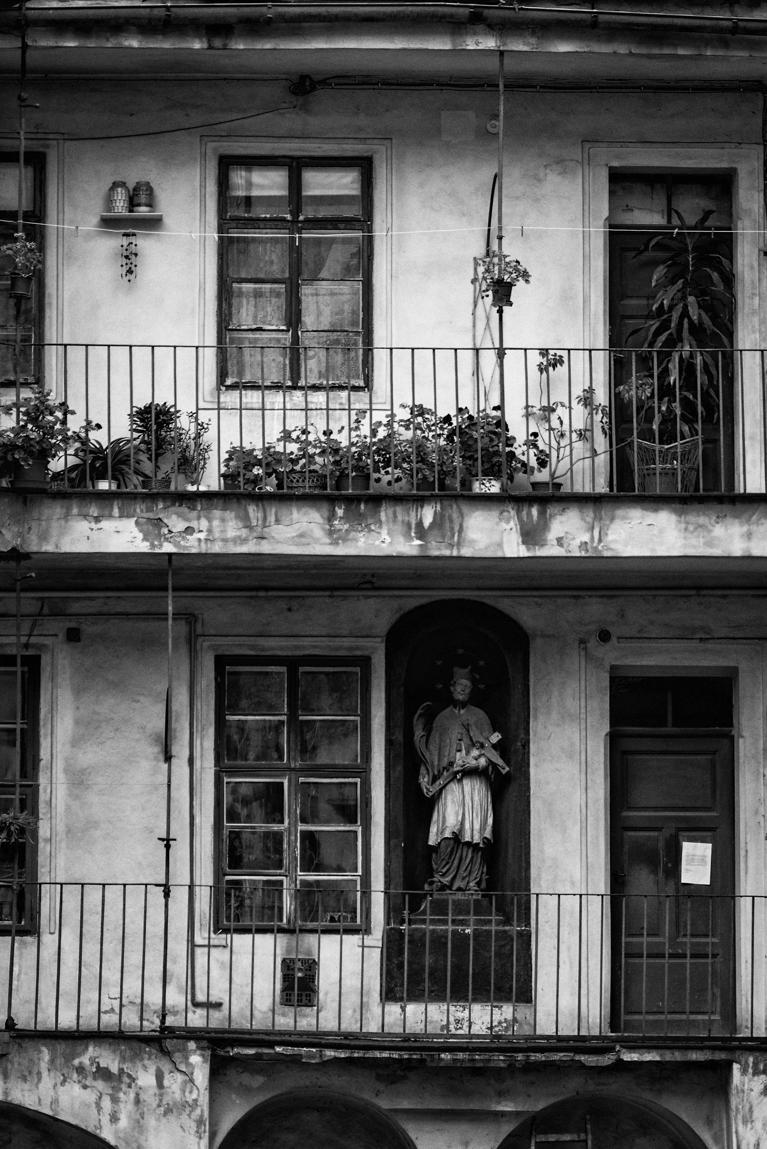 Black and white photo of an apartment building's second floor with potted plants on the balcony, two windows, a door, and a statue of a saint holding a book and a cross.