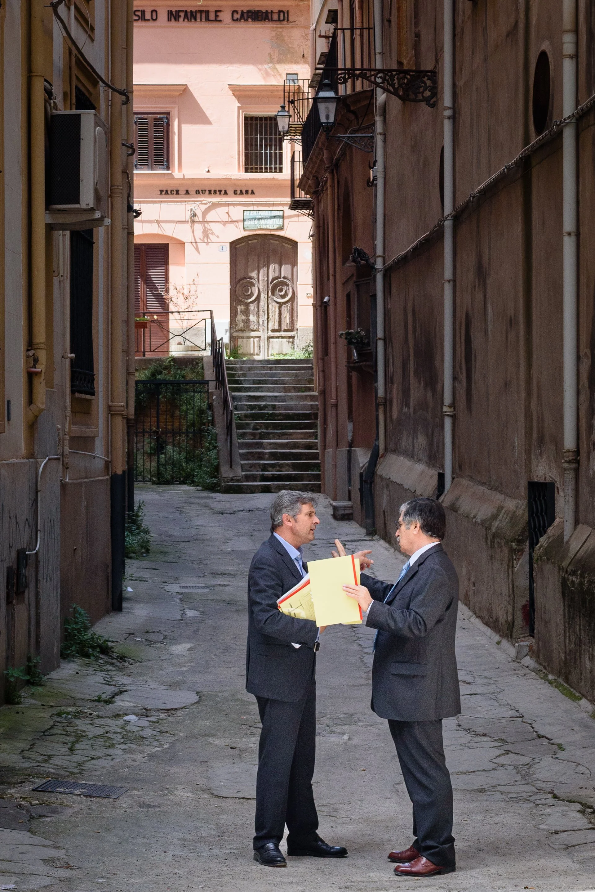 Two businessmen in suits having a discussion in a narrow, worn alleyway with cracked pavement, flanked by aged buildings with windows and balconies, and a staircase leading up to a pink building in the background.