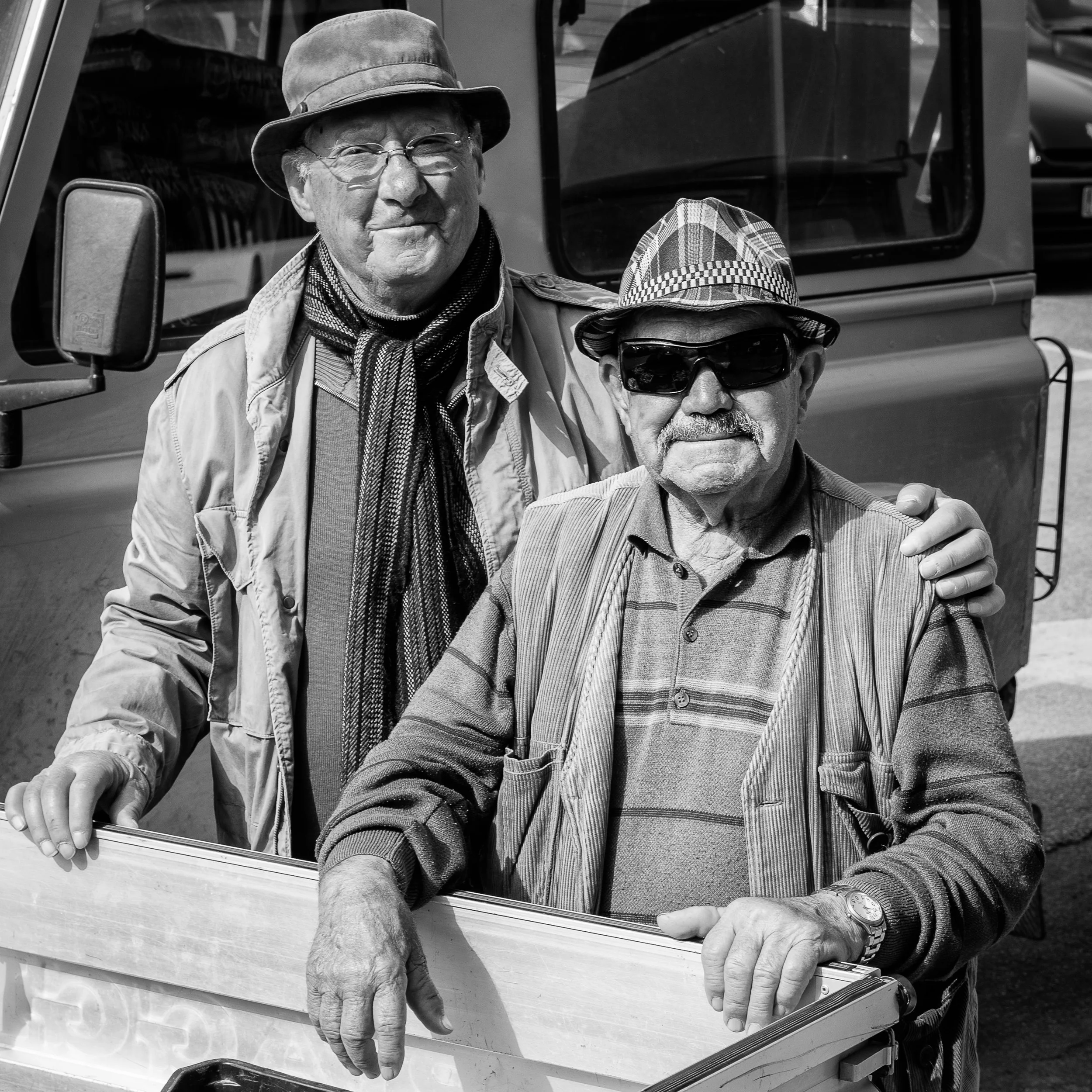 Two elderly men wearing hats and sunglasses standing outdoors near a vehicle, smiling. One man has his arm around the other.