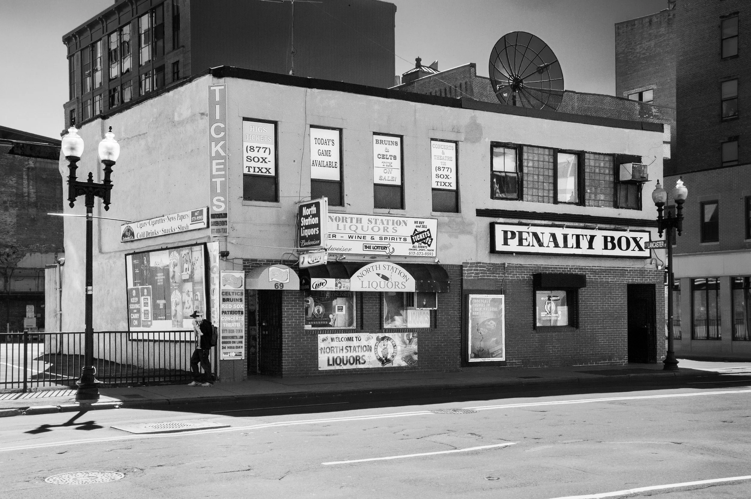 A black and white photo of a small corner liquor store and ticket booth with signs advertising tickets, tickets sales, and liquor. The building has windows, signage, and a fire escape, with a person walking on the sidewalk.