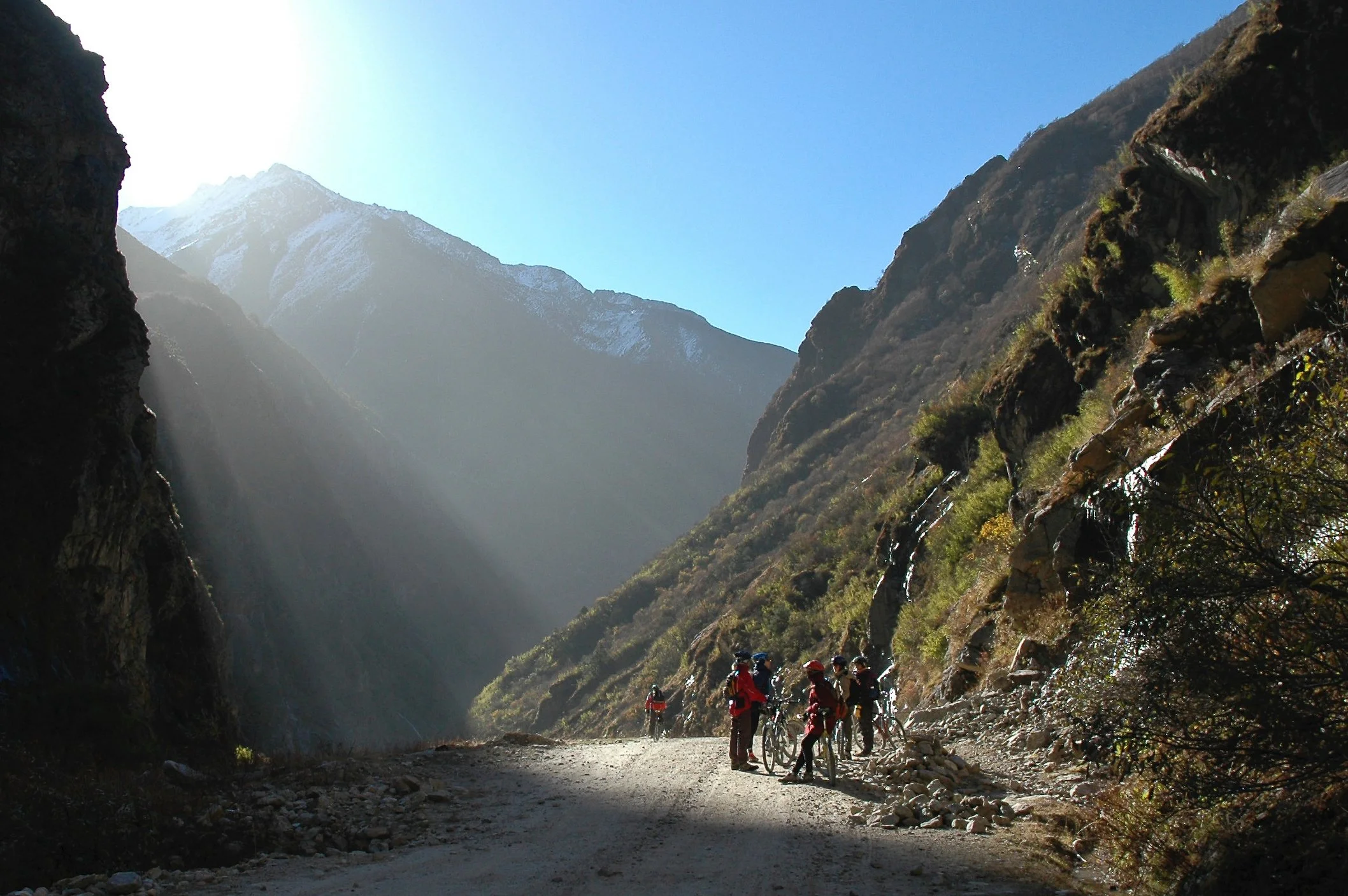 A group of hikers with backpacks and bicycles on a mountain trail surrounded by tall rocky cliffs and snow-capped peaks in the background.