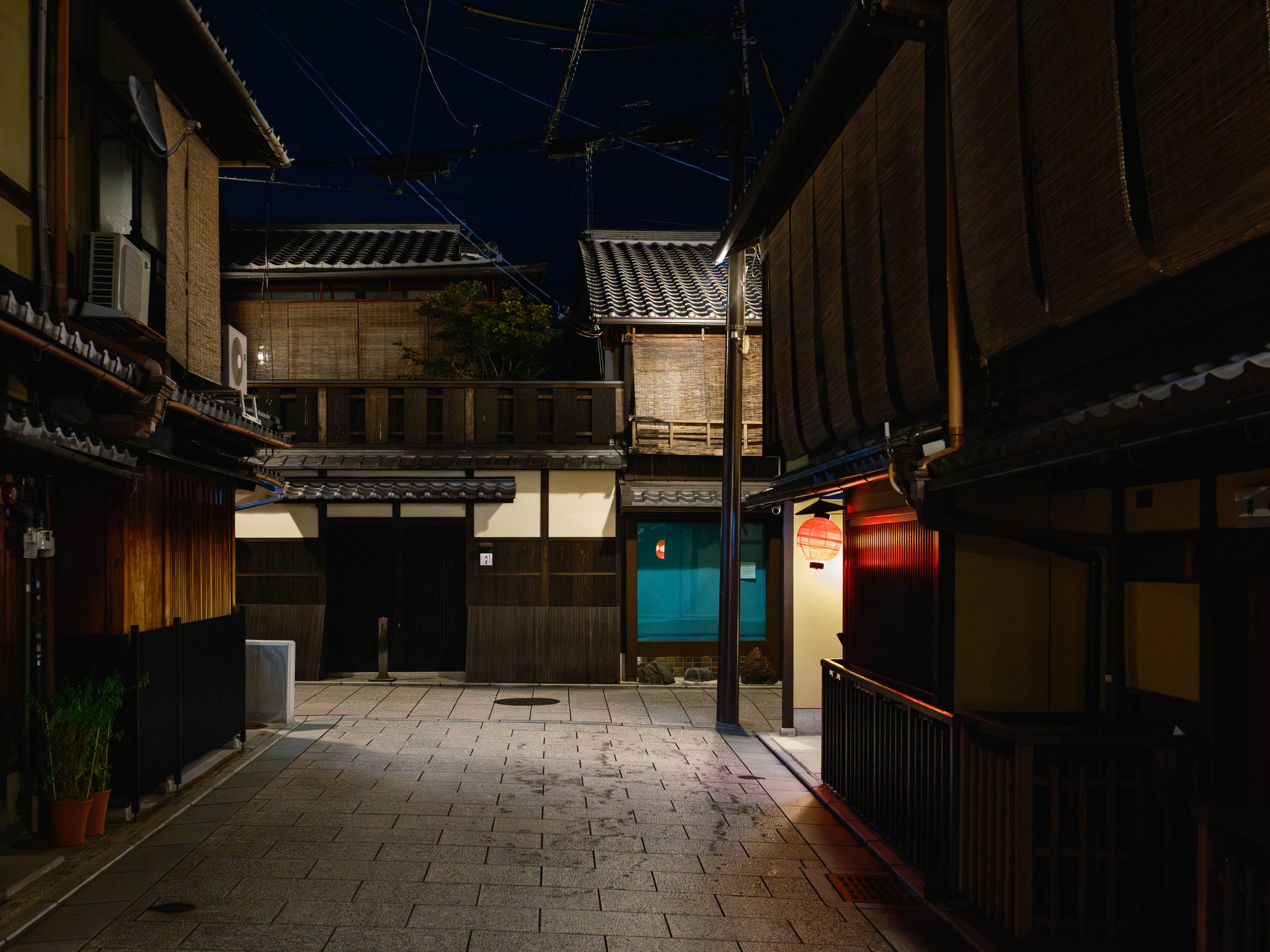 A quiet traditional Japanese alleyway at night, with wooden buildings, lanterns, and tiled roofs.