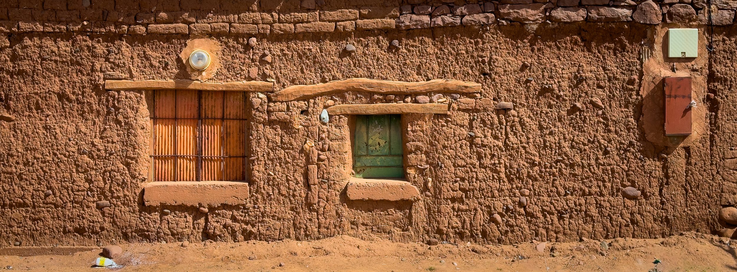 Close-up of an adobe wall with two small windows, one barred with rusted metal and one painted green, and surrounding electrical boxes and a light fixture.