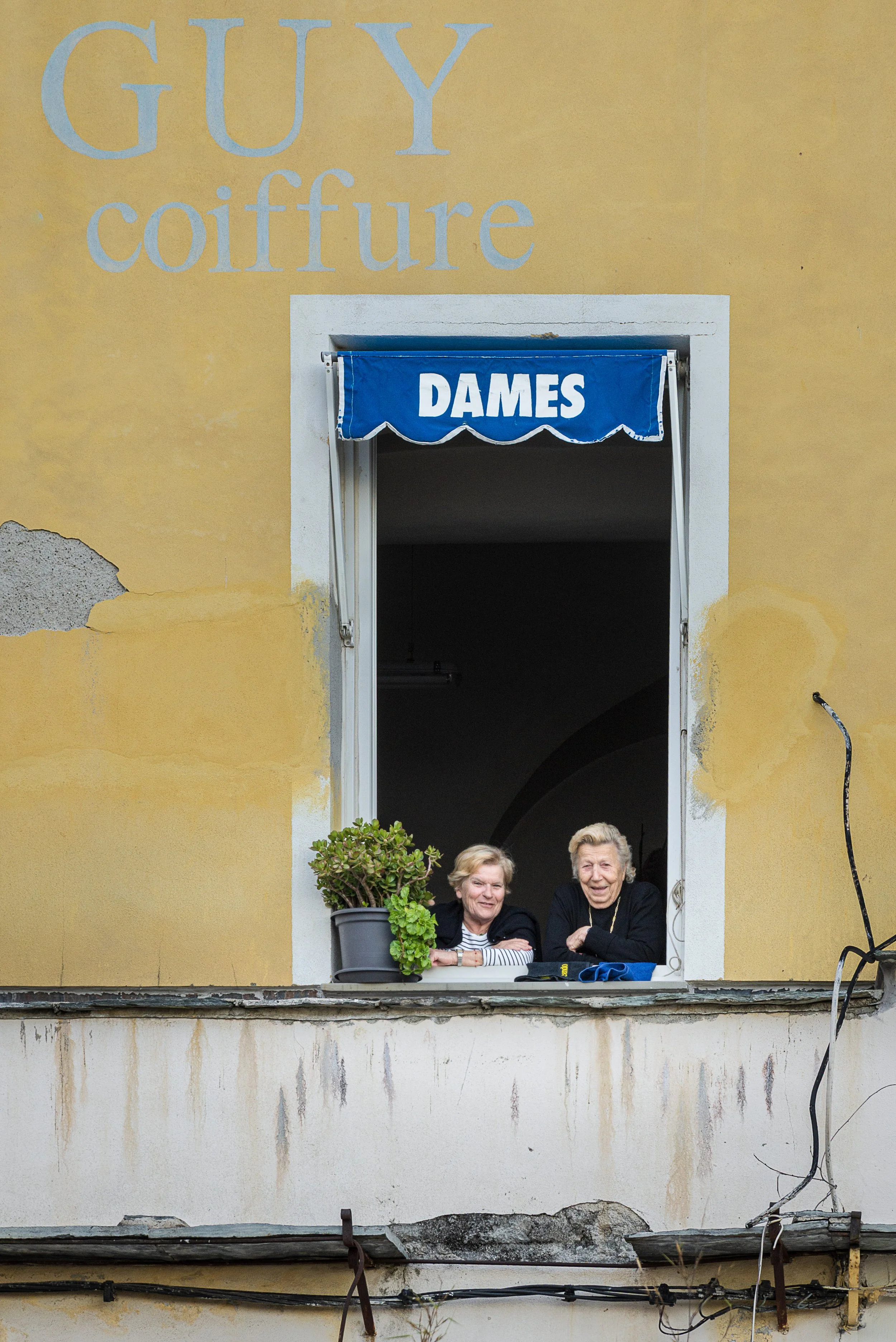 Two elderly women leaning out of a window with a blue awning labeled 'DAMES', on a yellow building with peeling paint and exposed wiring.