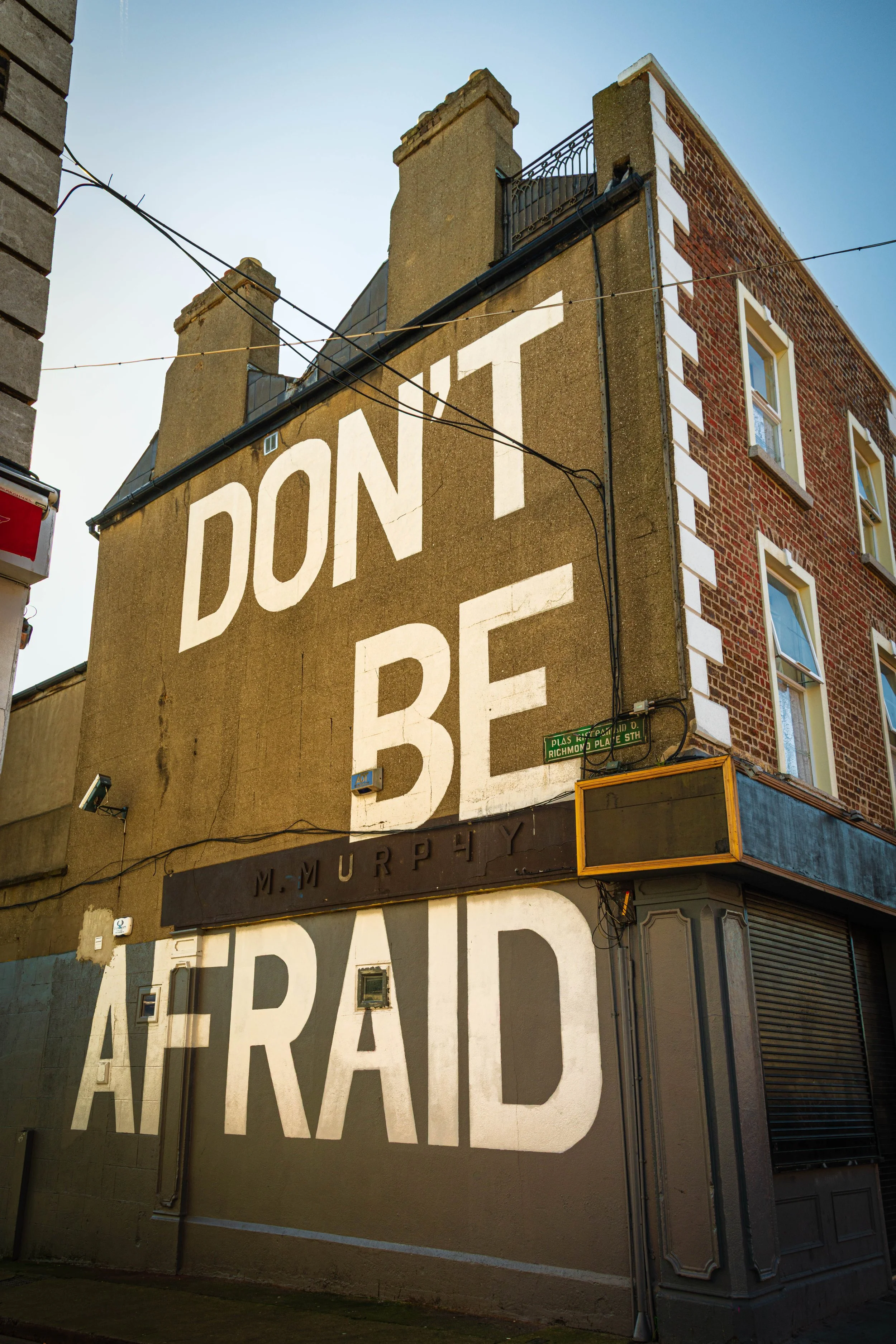 A large mural on the side of a building reads 'DON'T BE AFRAID' in white letters on a brown background. The building is made of red brick and has several windows, with a street sign indicating the intersection of Plas Street and Richmond Place SW.
