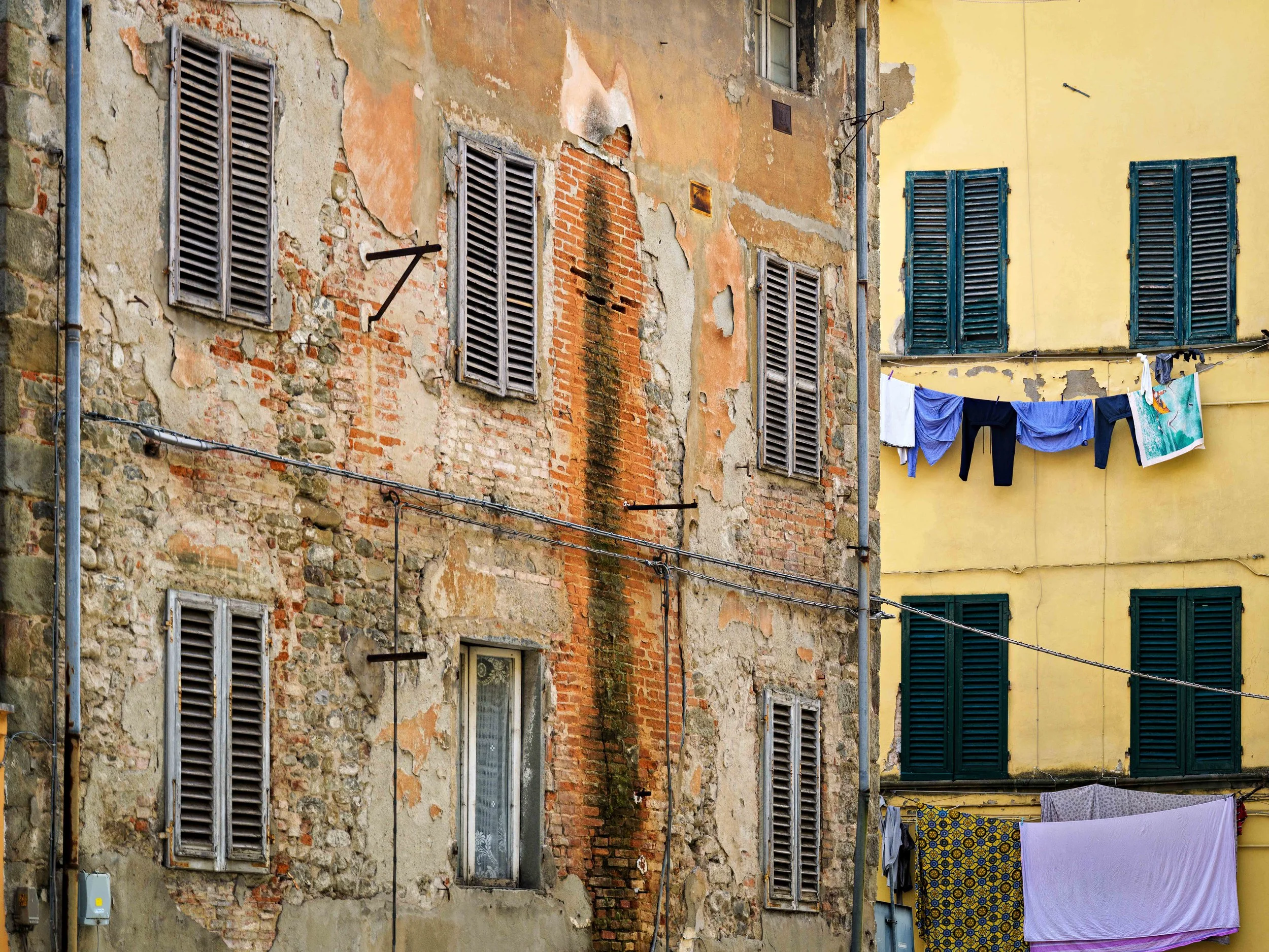 Exterior wall of two old buildings with peeling paint, shutters, and laundry hanging on a clothesline.