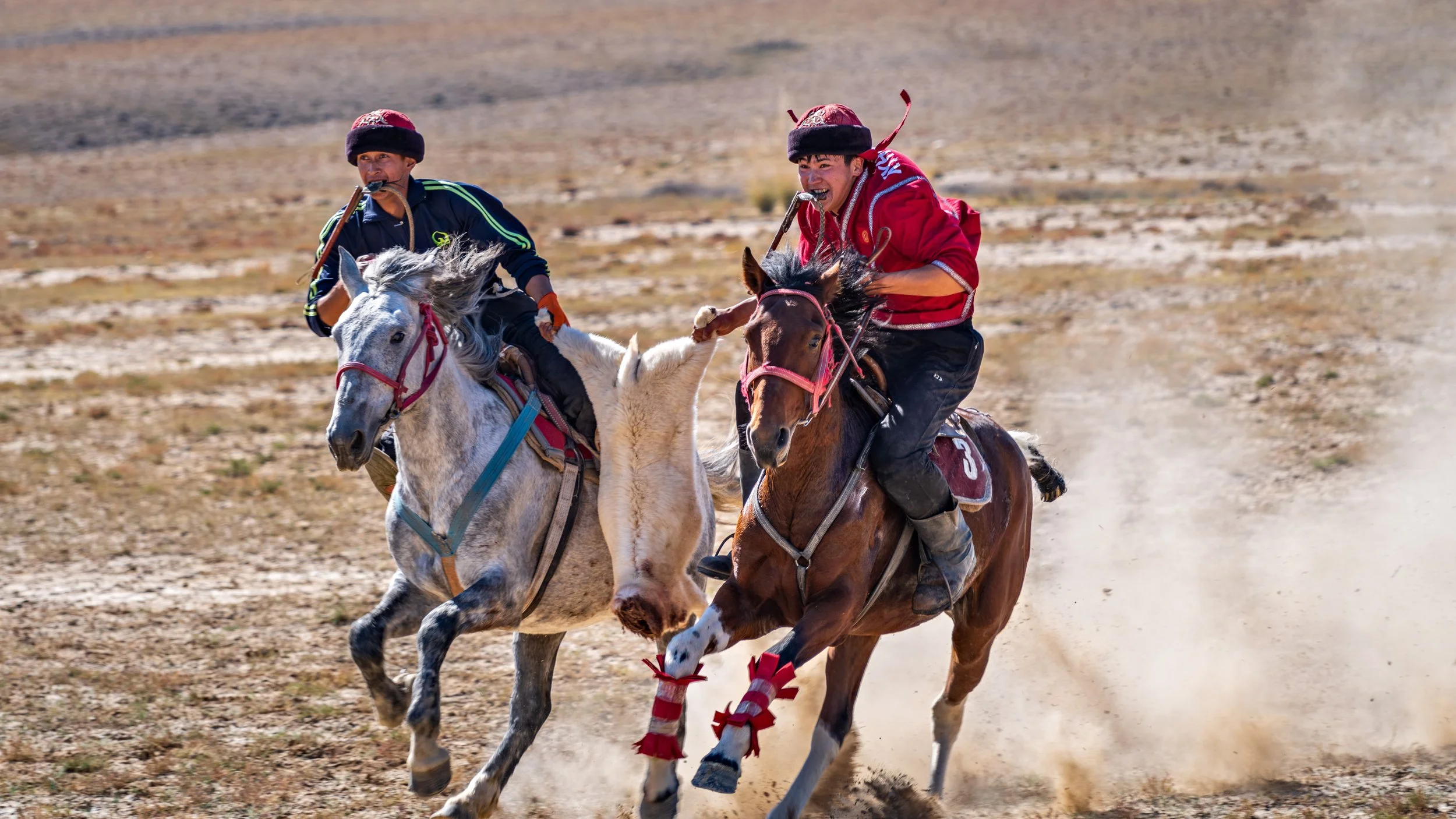 Two men riding horses with a goat jumping between them in a dry, open landscape