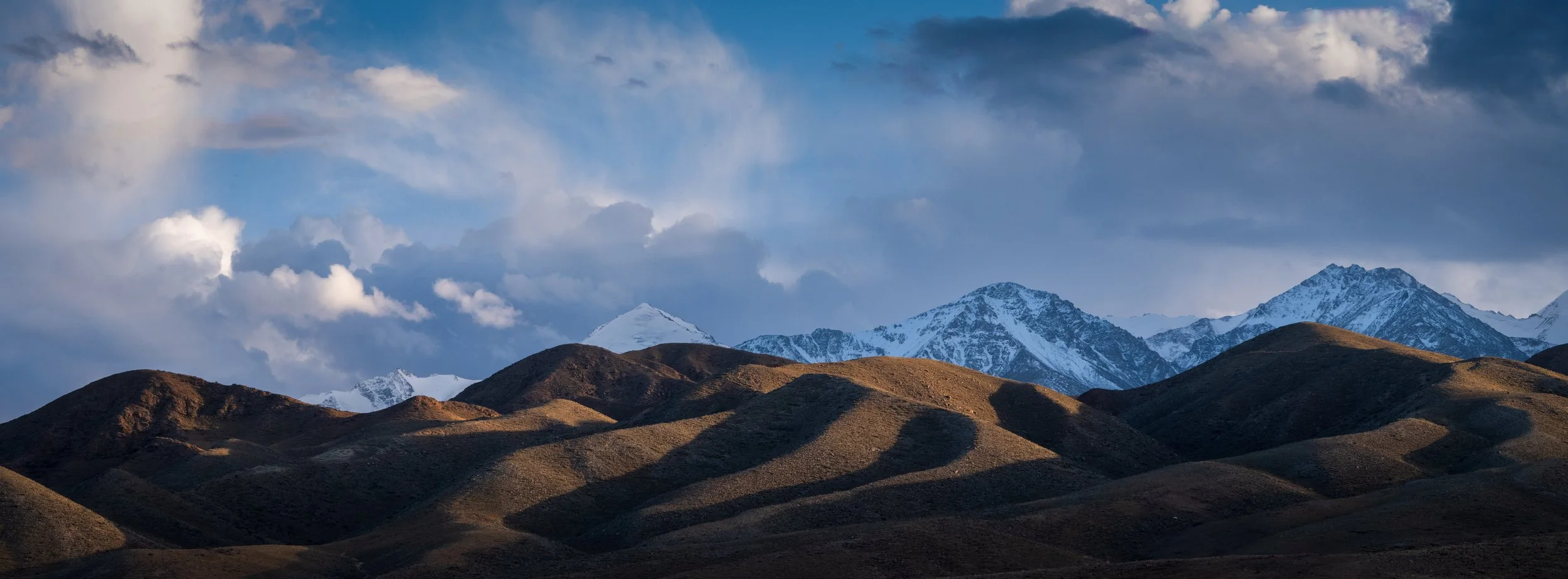 Scenic view of snow-capped mountains in the background with brown rolling hills in the foreground under a partly cloudy sky.