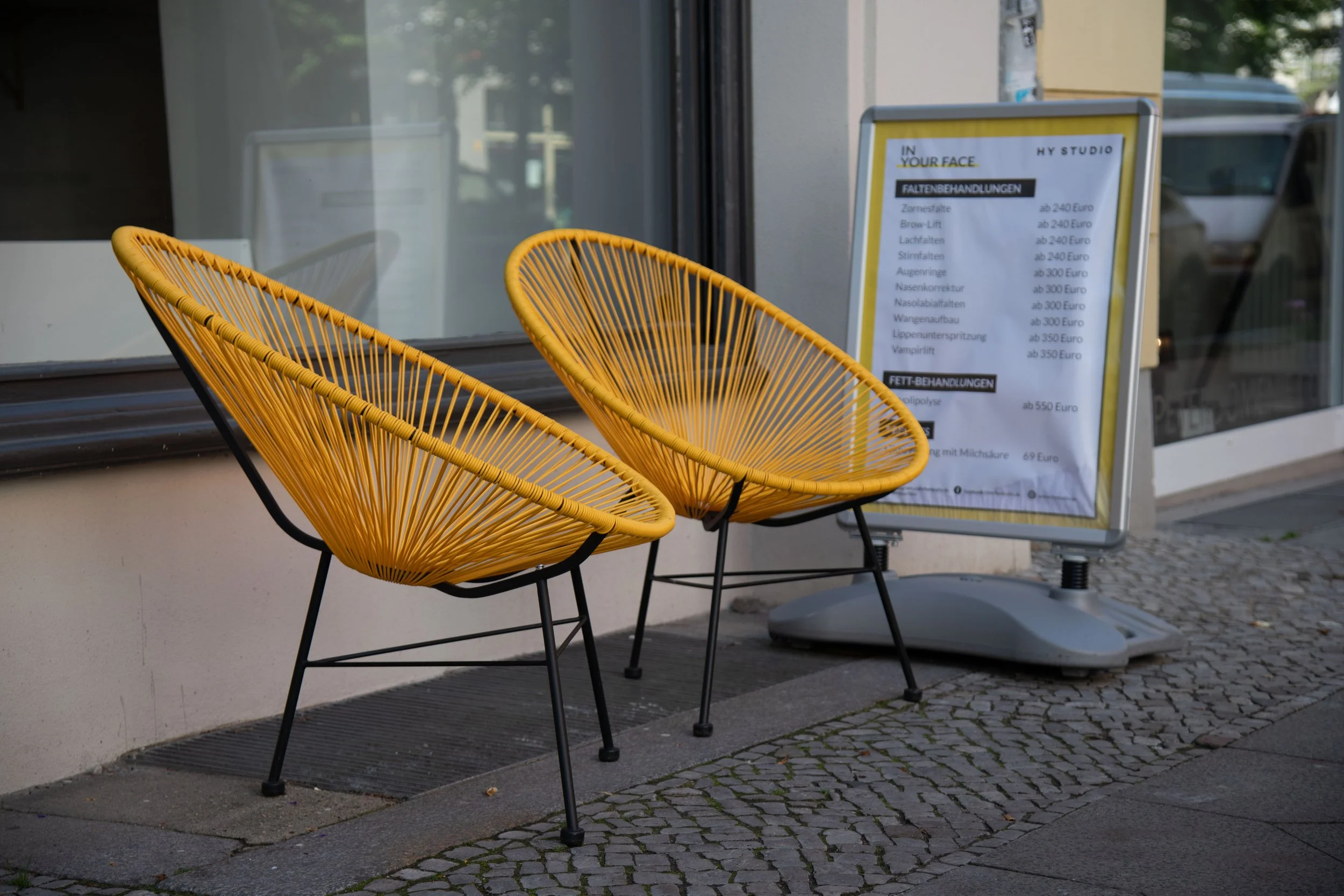 Two yellow outdoor chairs with black metal legs in front of a storefront window and a white sandwich board sign with a menu.