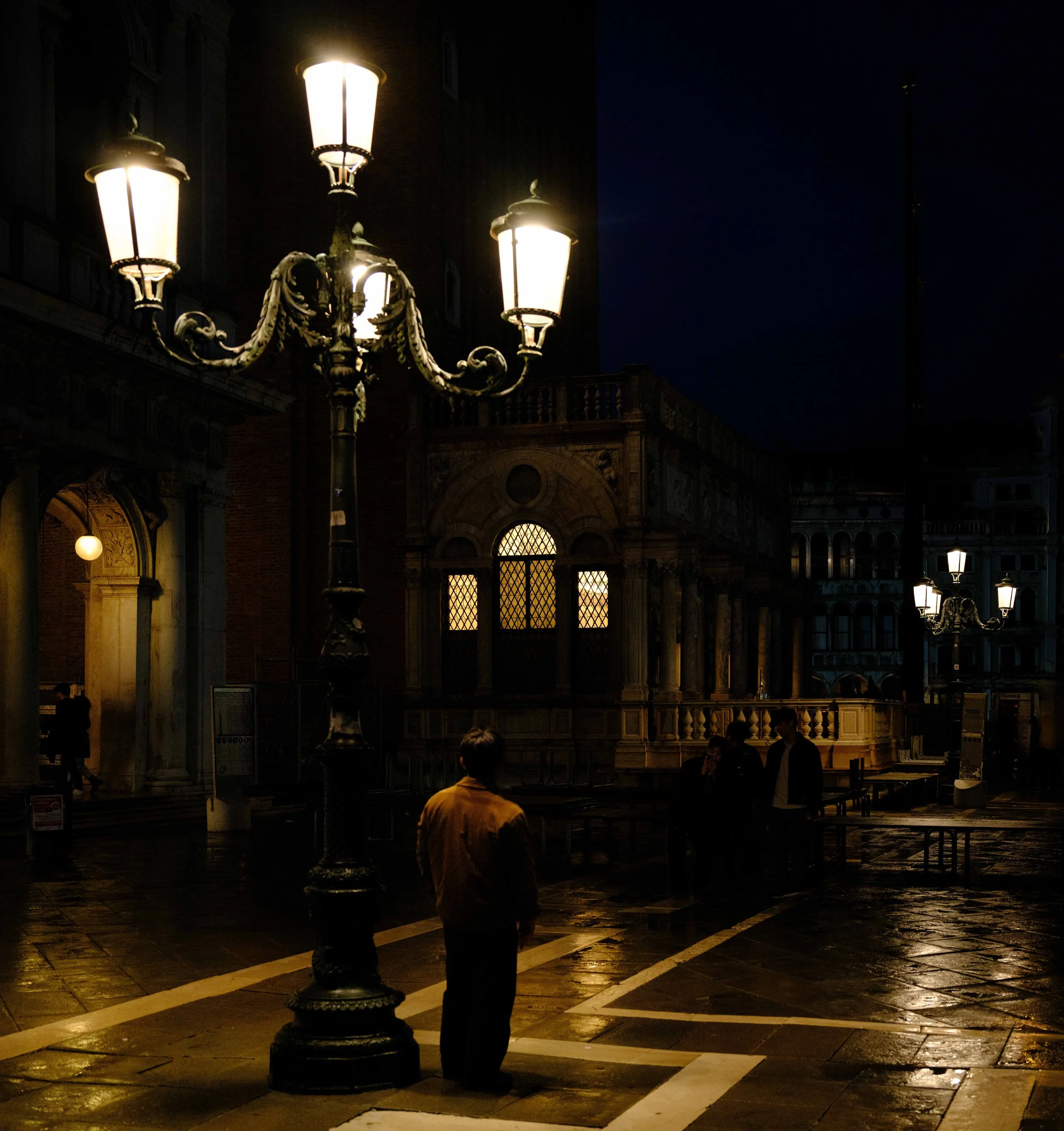 Night scene of a wet city street illuminated by vintage street lamps, with a man in a yellow jacket standing under the lamps, and a group of people further away near a historic building with ornate windows.
