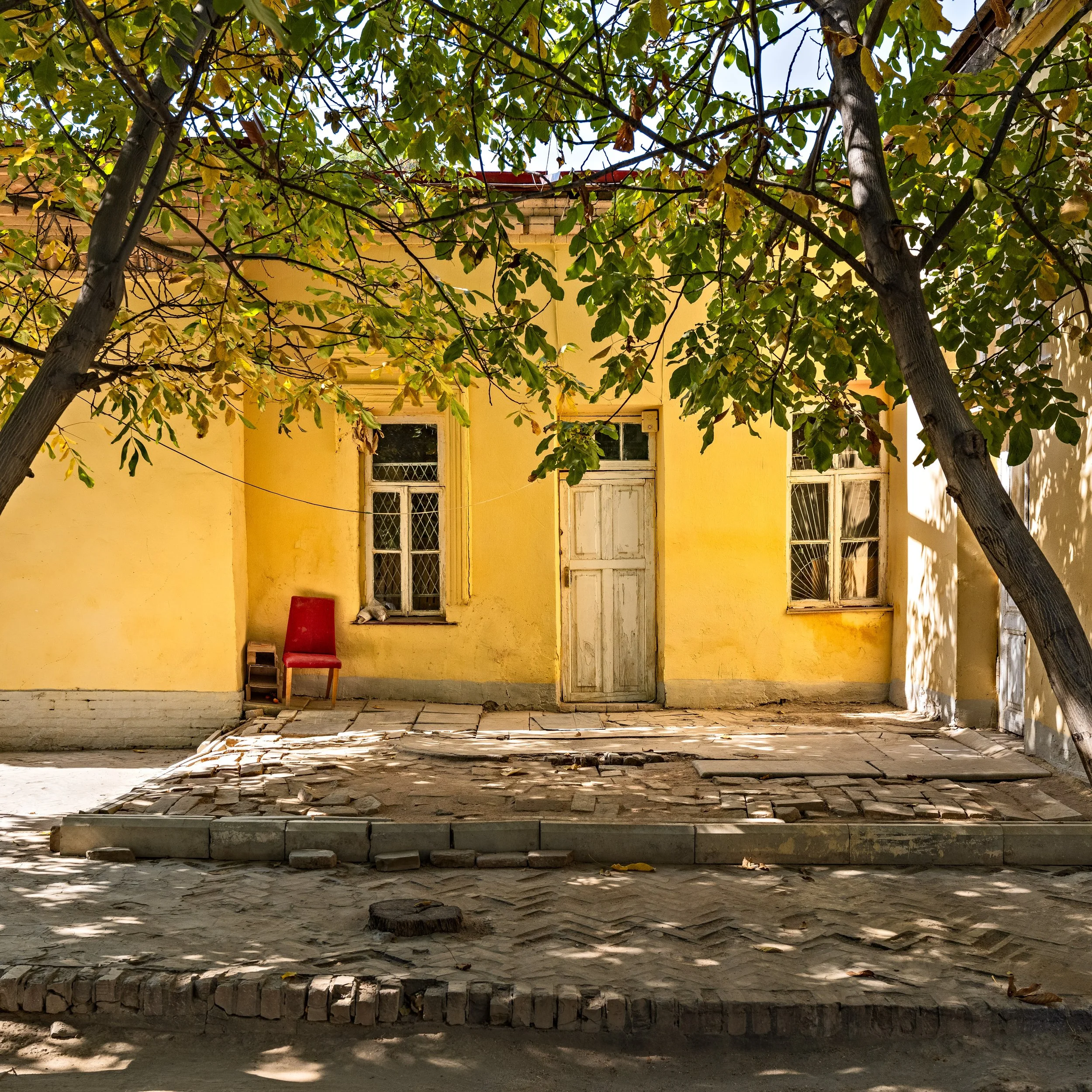 A small, yellow house with three windows, a white door, and a red chair placed beside the left window. Two large trees with green leaves partially shade the house in an outdoor setting.