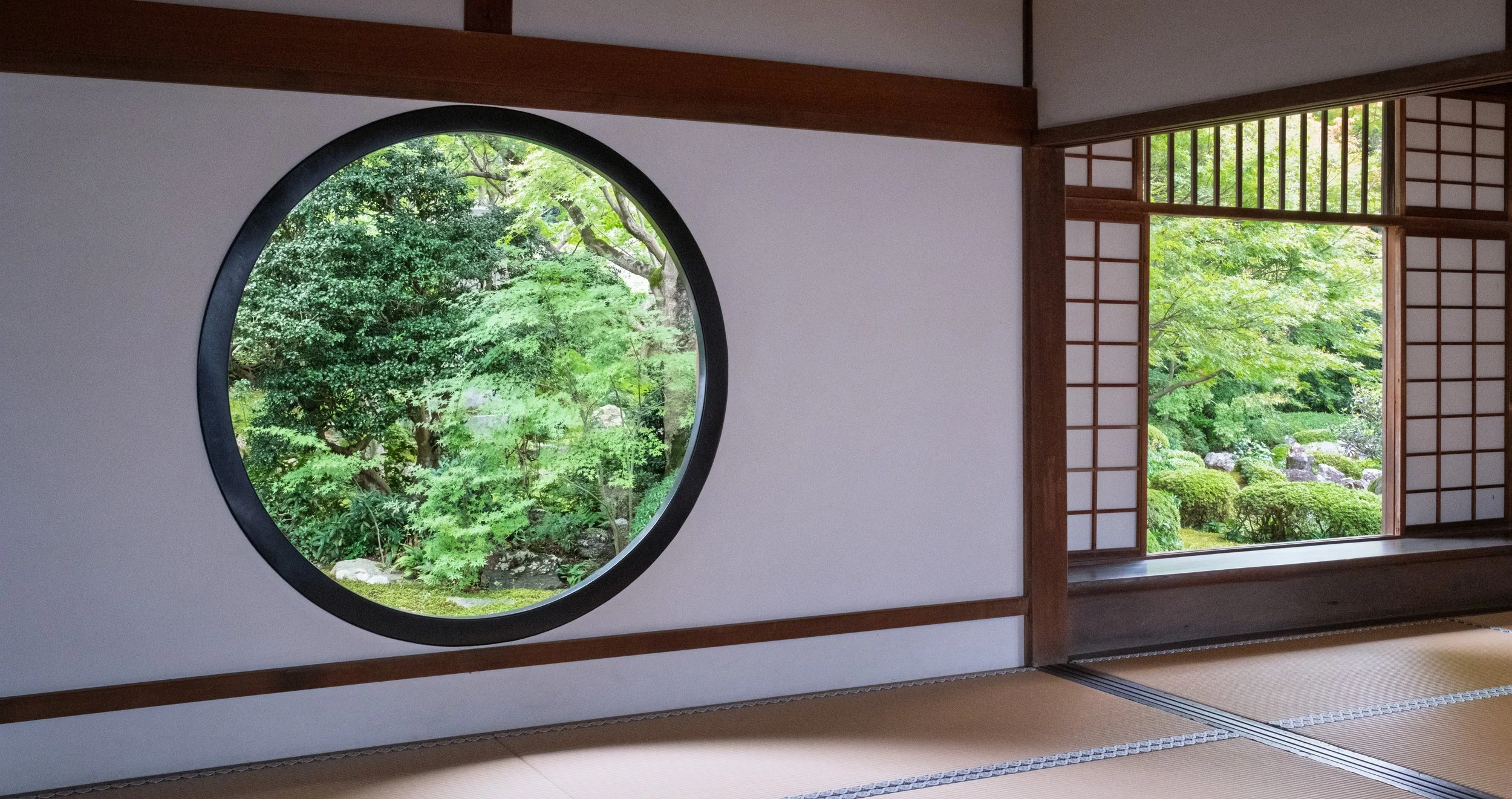 View through round window and sliding shoji doors showing a lush Japanese garden with green trees and bushes.