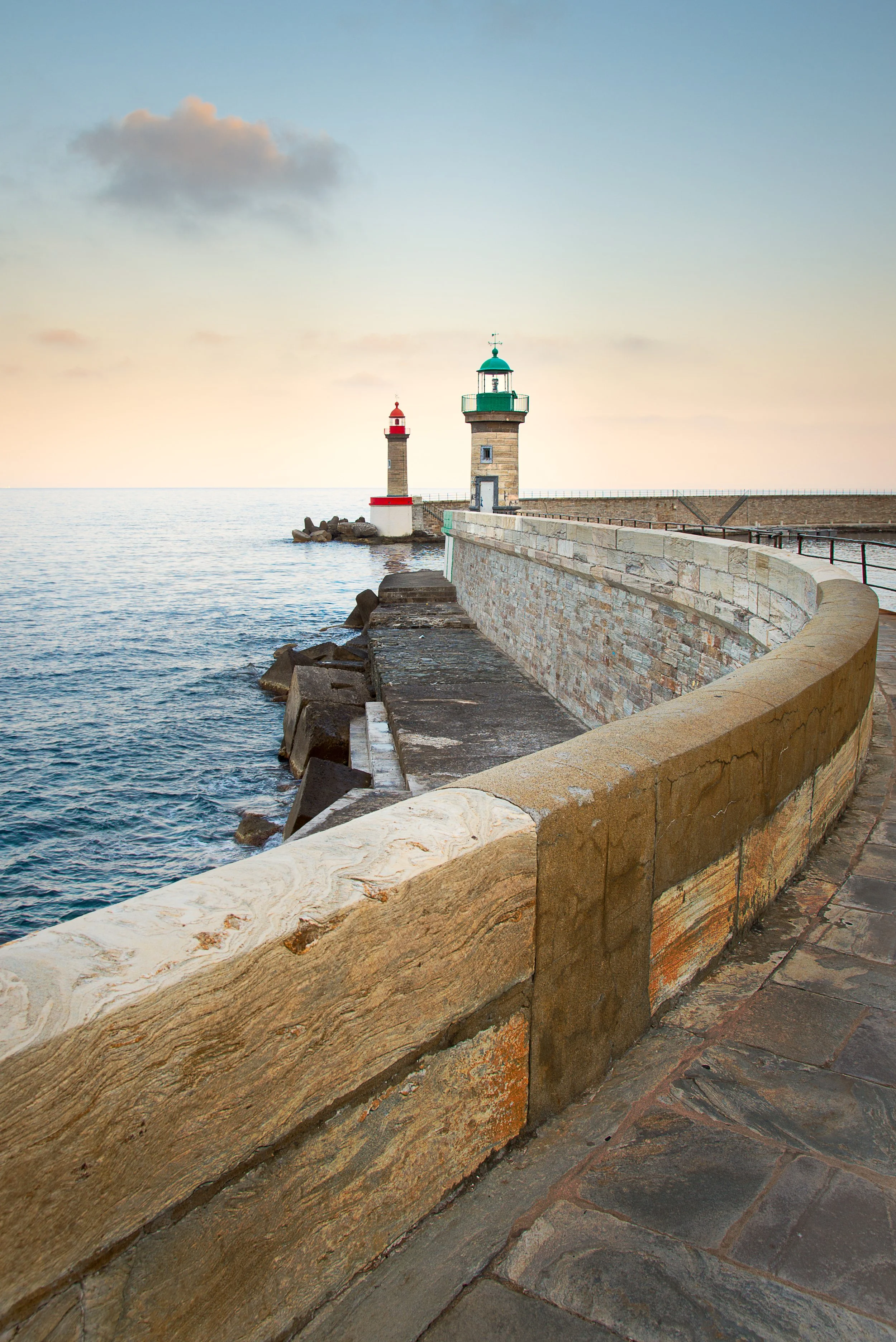 Two lighthouses along a rocky shoreline at sunset, with an arched stone promenade in the foreground.