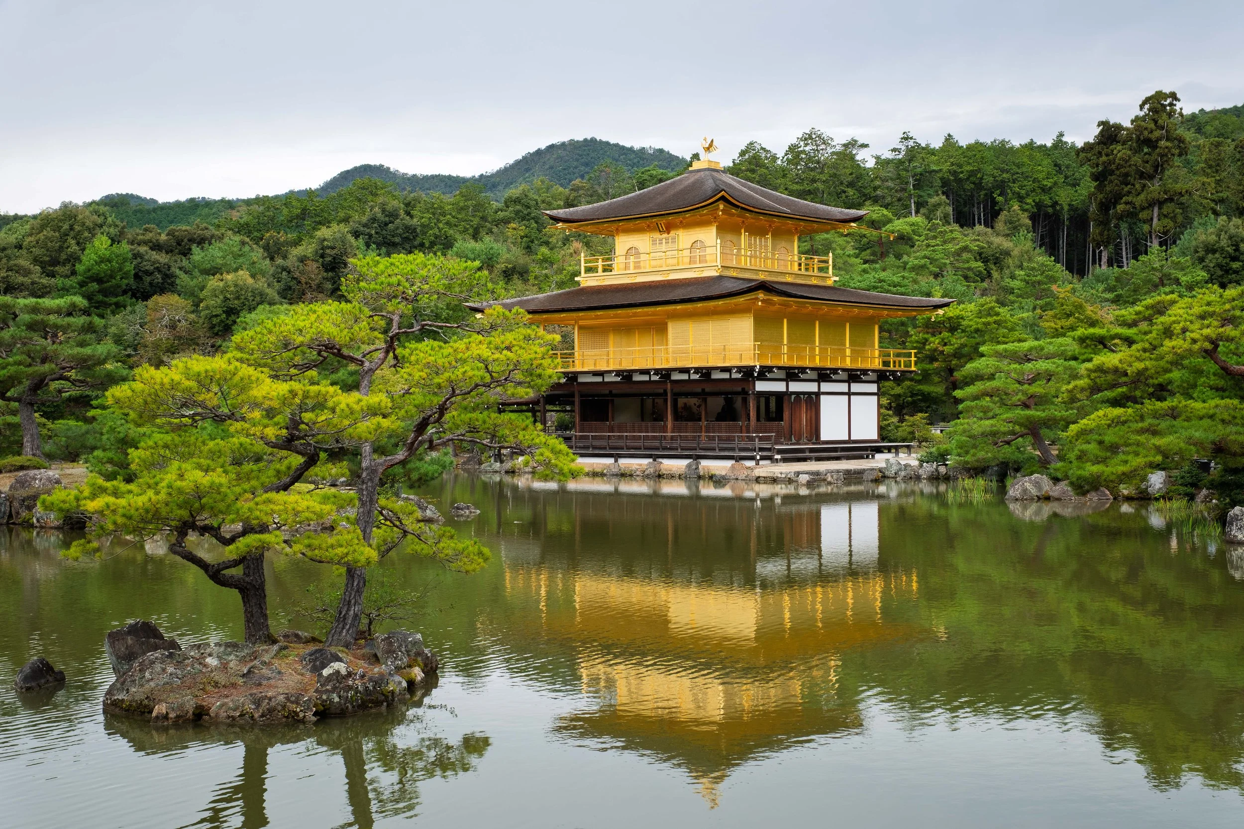 Traditional Japanese temple with golden exterior and layered roof, situated beside a pond with green trees and mountain in the background.