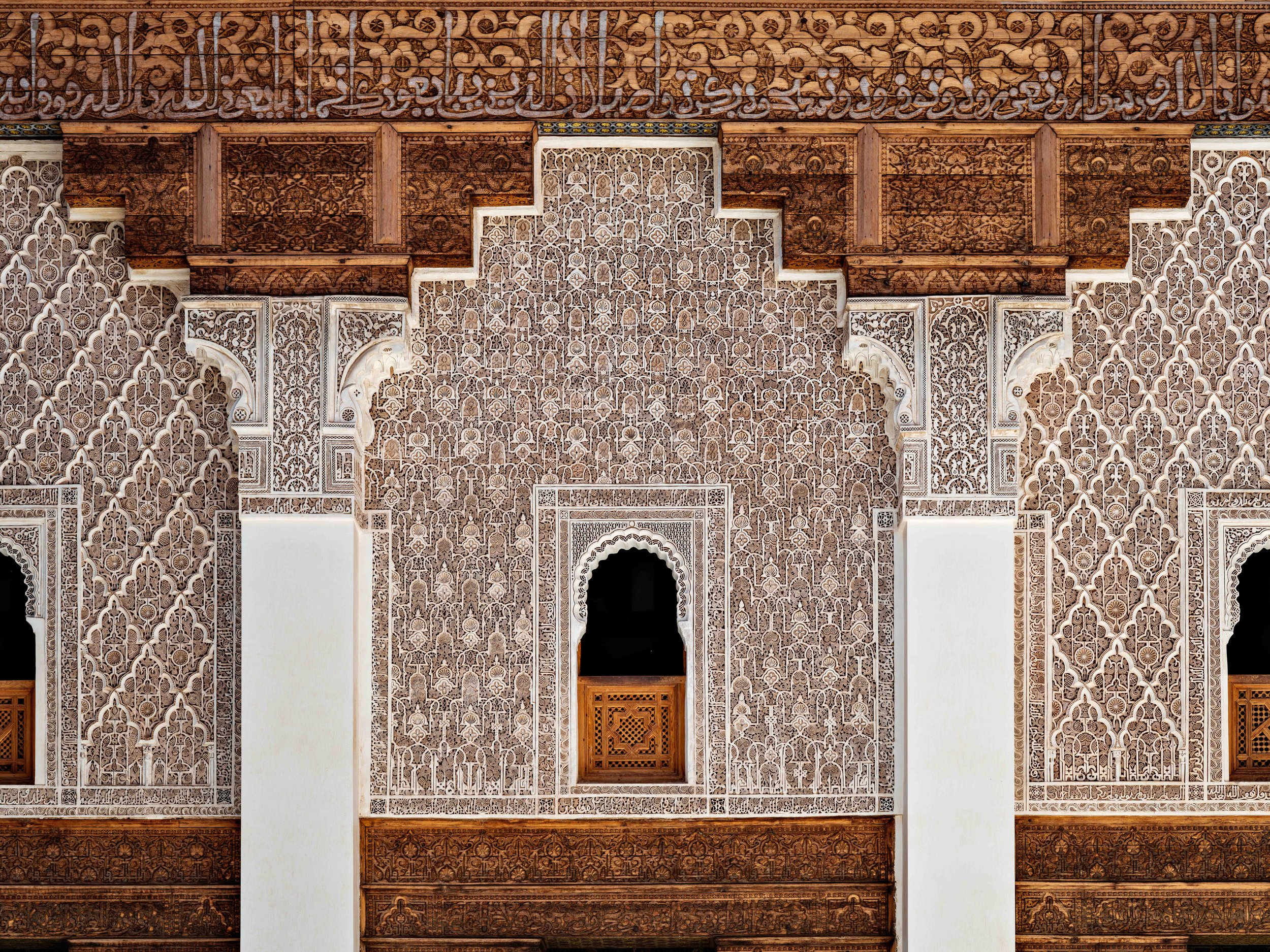 Intricate Moroccan-style wall with detailed wood and carved white plaster patterns, featuring central arched window, surrounded by ornate geometric and floral designs.