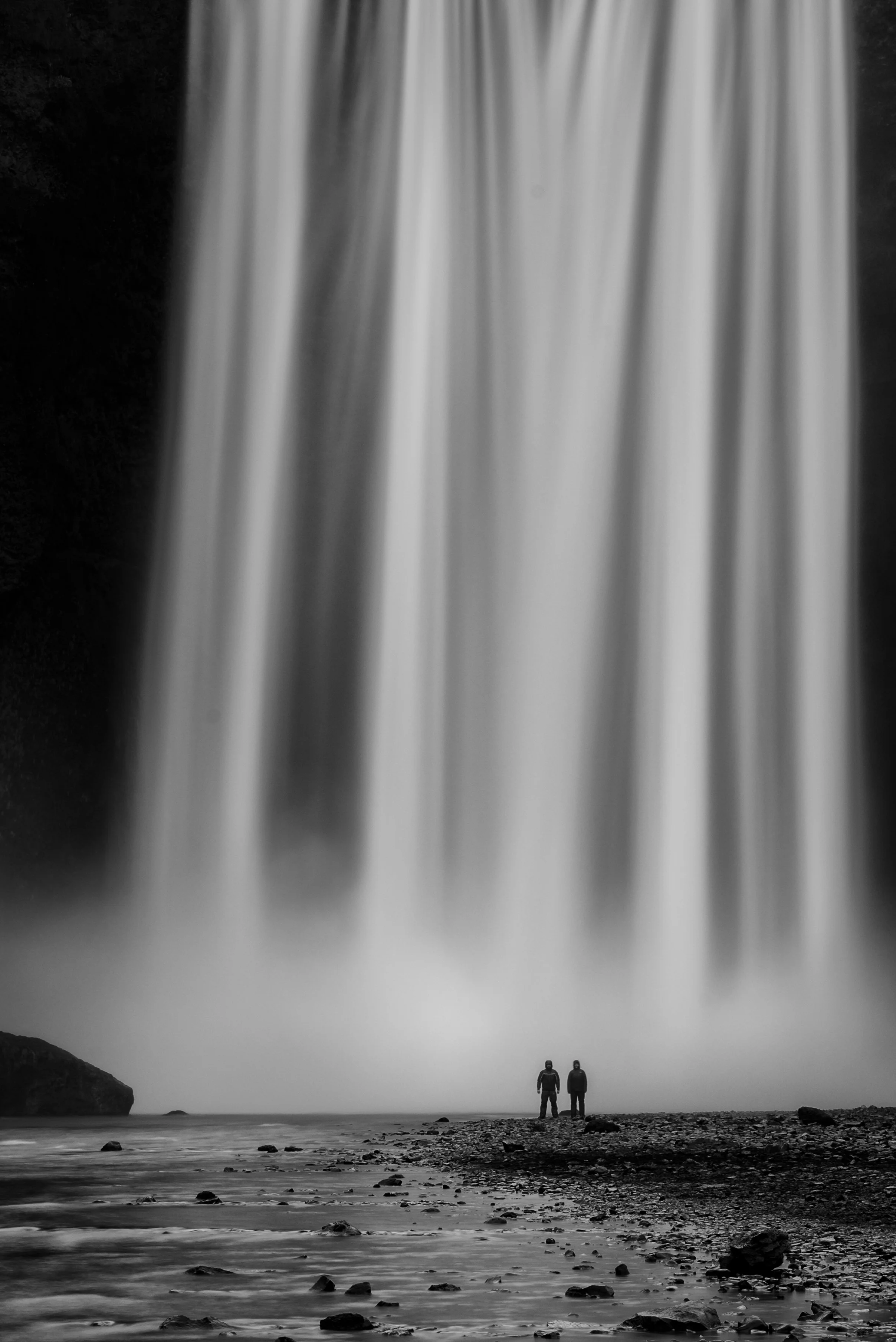 Two people standing on rocky shoreline near waterfall in black and white.