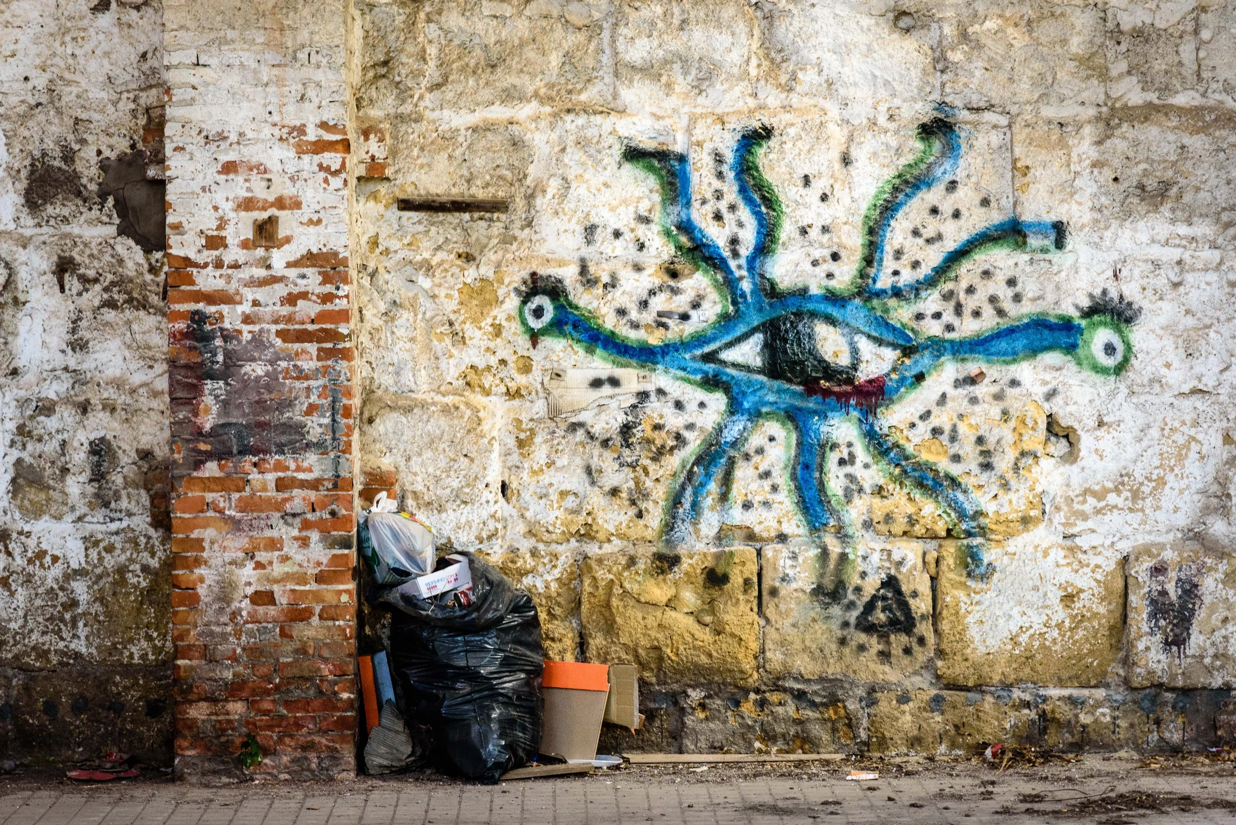 Graffiti art of a blue eye with green and blue tentacle-like extensions on an aged, damaged brick and stone wall, with garbage bags and debris on the ground.