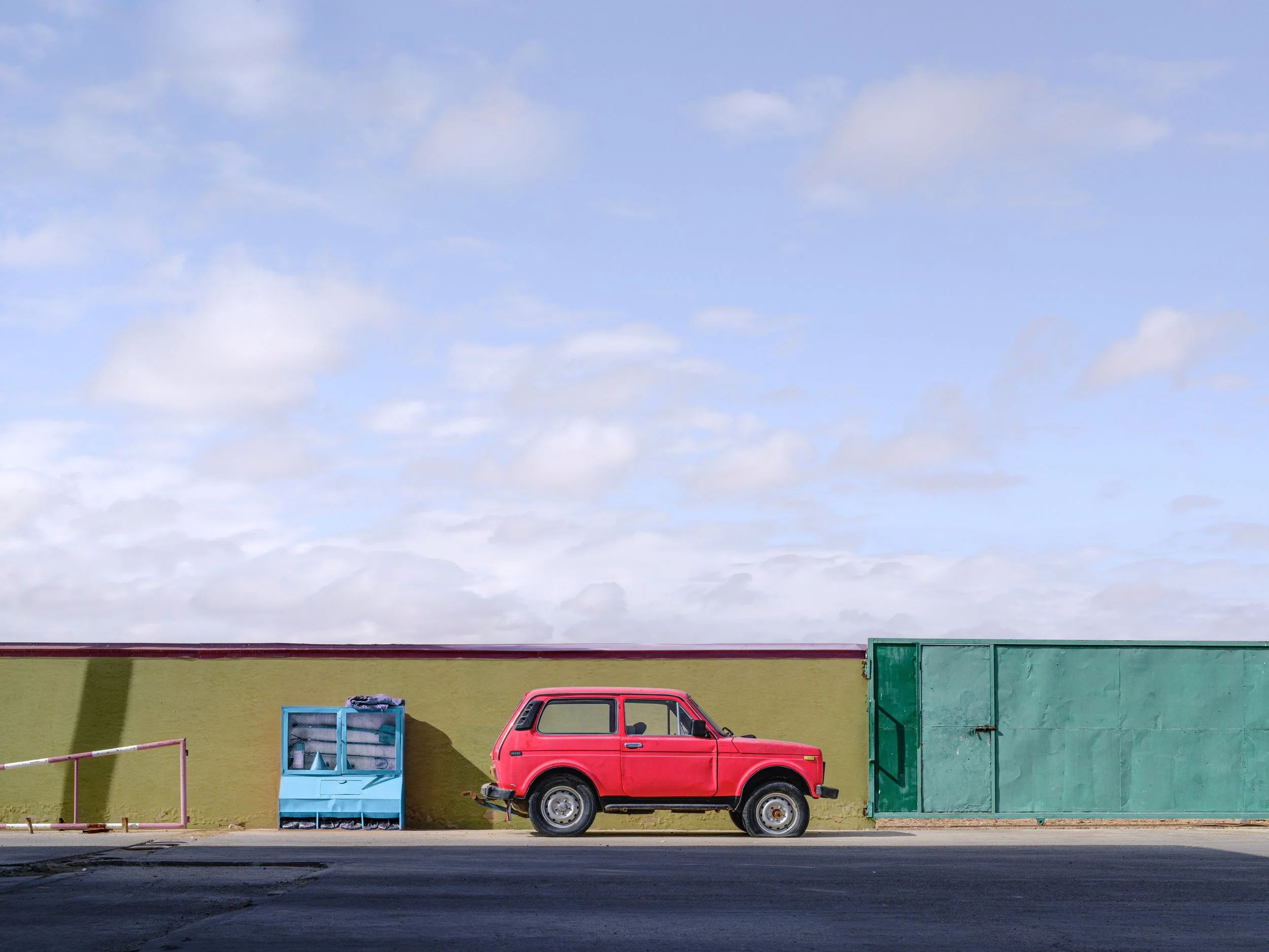A red compact SUV parked in front of a green wall with a blue vending machine and a large green gate, under a partly cloudy sky.