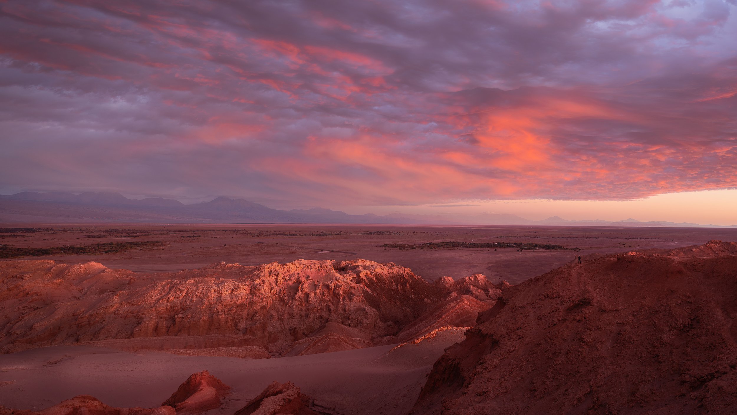 Sunset over a desert with reddish rock formations and a vast flat plain, with a colorful sky filled with purple and pink clouds.