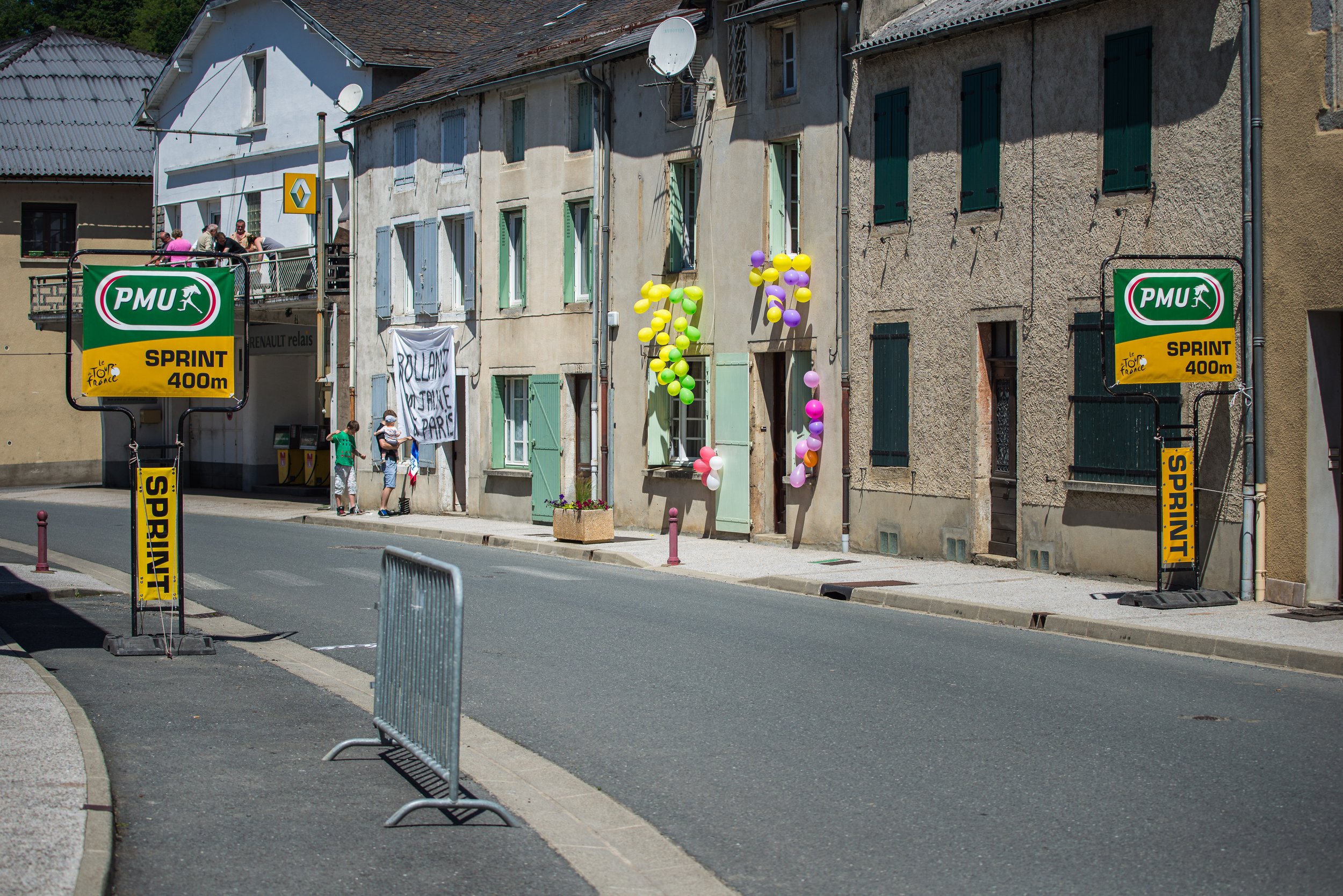 Street scene in a town with balloons attached to the wall, children and adults standing on the sidewalk, and signs indicating a 400-meter sprint for a cycling race. Colorful balloons, a banner, and nearby buildings are visible.
