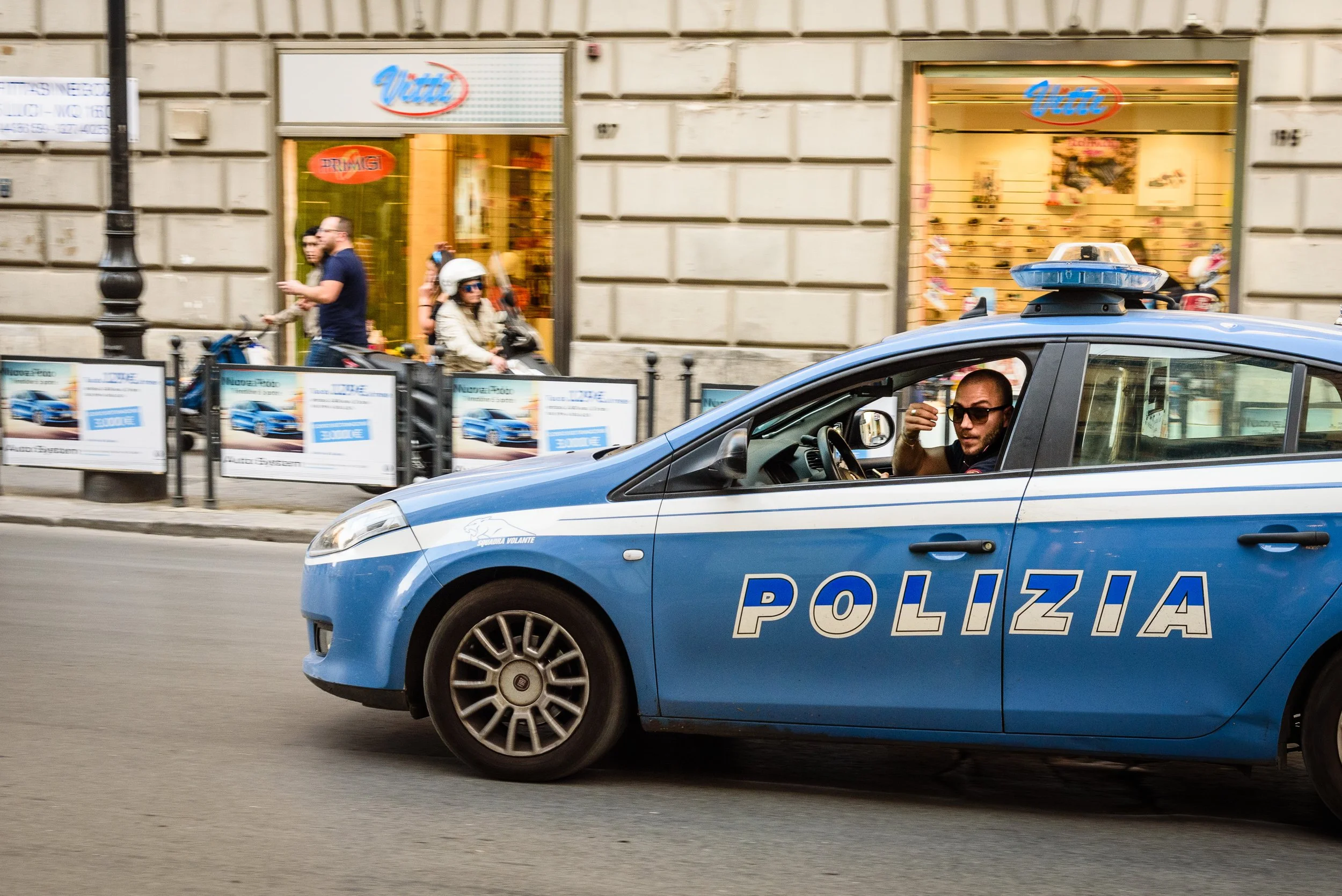 Italian police car with officer inside, passing by pedestrians and scooter riders on a city street with shopfronts.