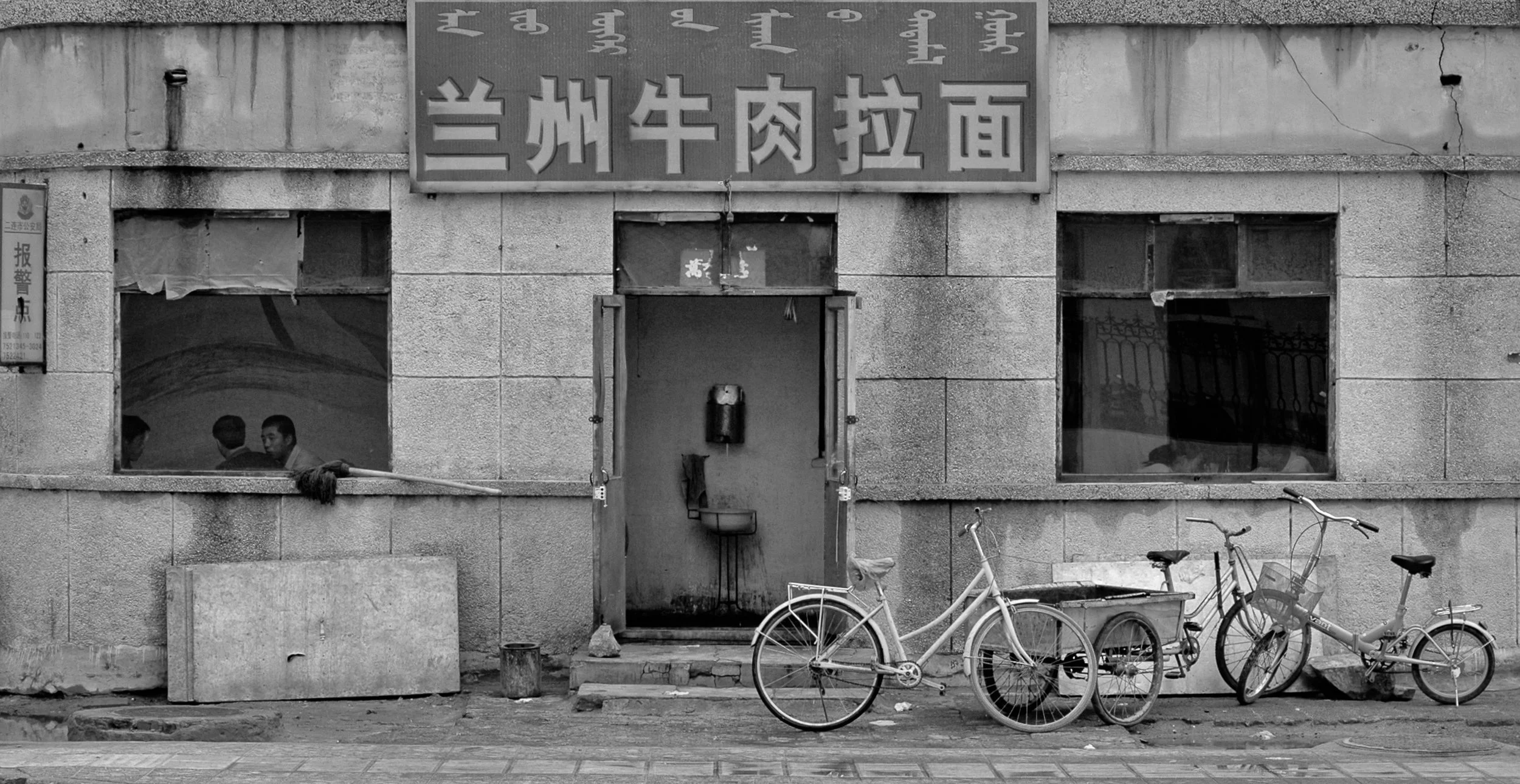 Black and white photo of the exterior of a building with a sign in Chinese characters, two bicycles parked in front, and three people visible through a window inside.
