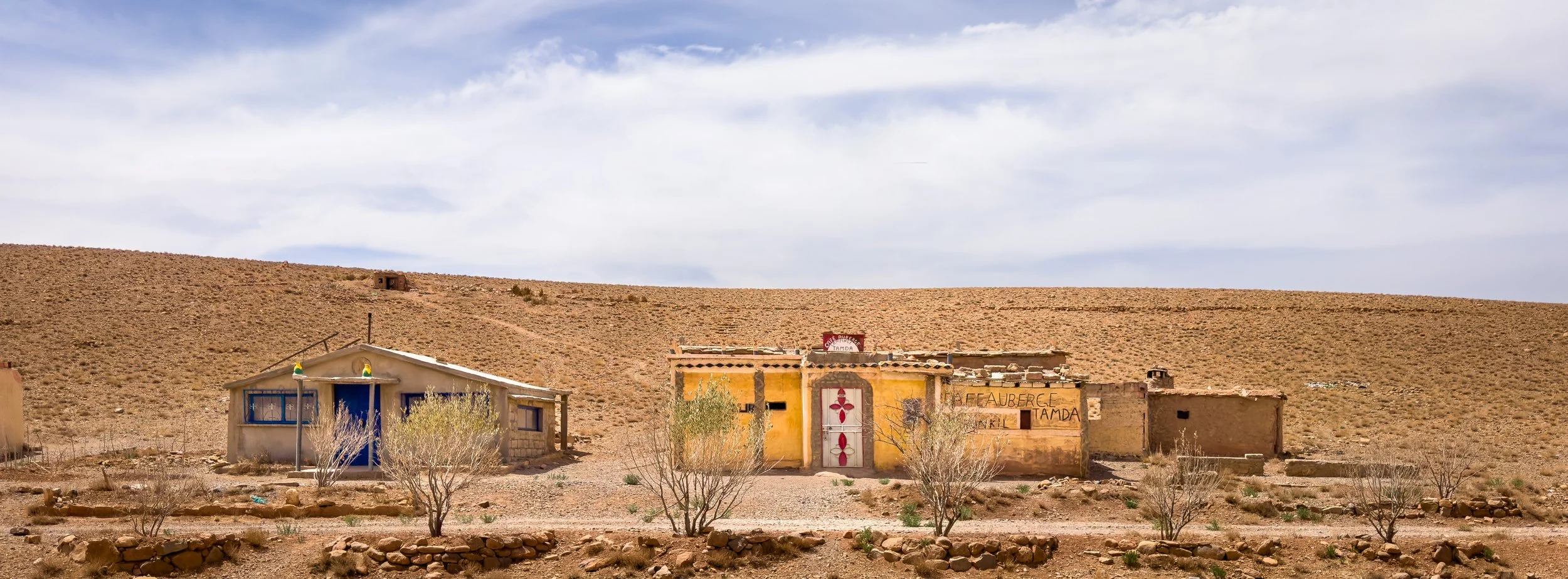 A row of small, colorful buildings in a barren desert landscape with a hill in the background and a cloudy sky overhead.